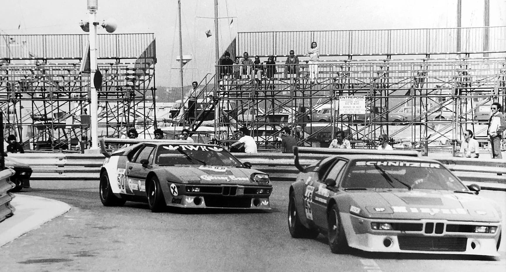 A black and white photograph of two Procars at Monaco's swimming-pool section, one lettered TWR and one SCHNITZER, with grandstands and onlookers behind.