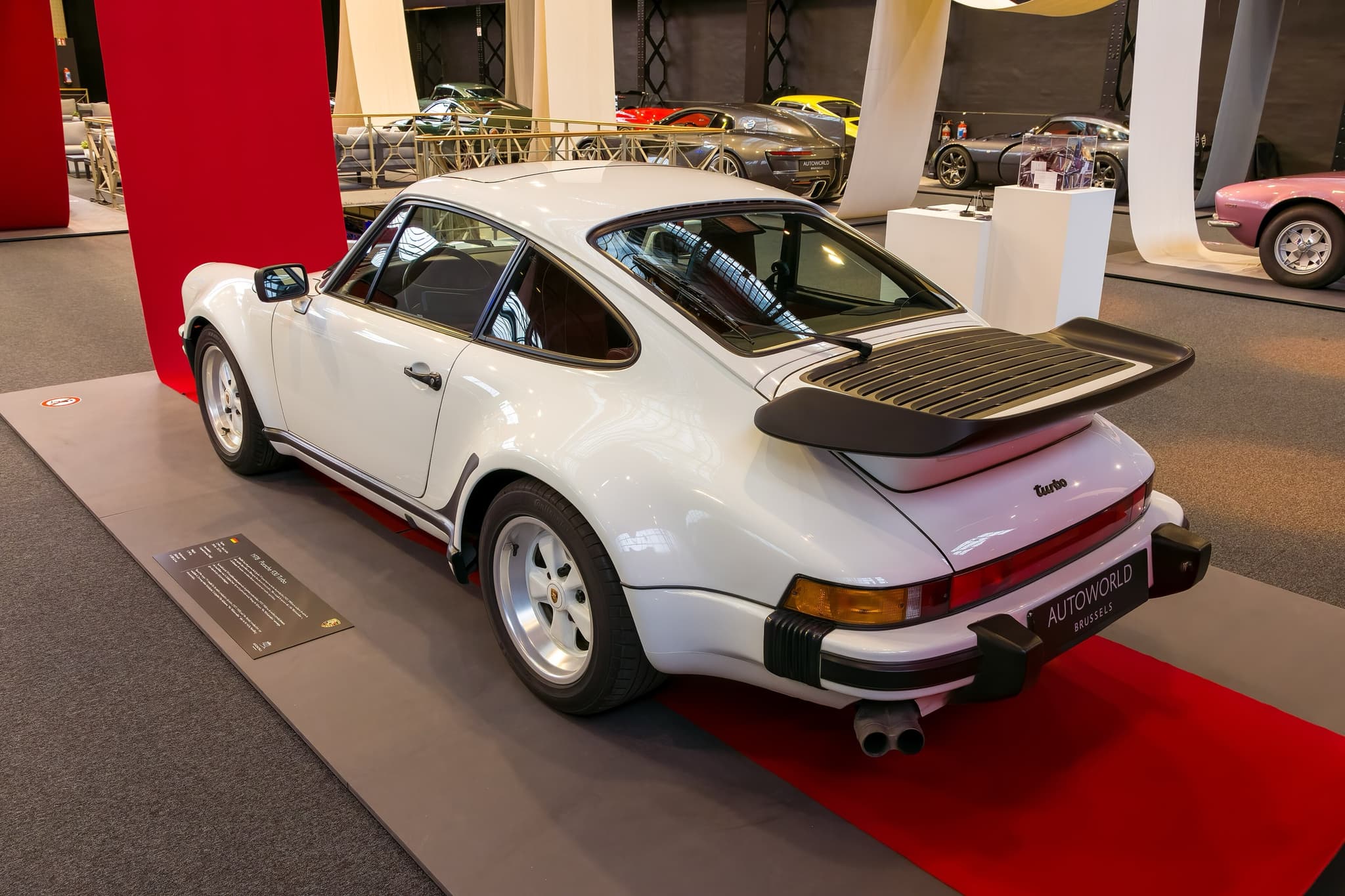 A white Porsche 911 Turbo with a whale-tail spoiler on a red-carpeted museum stand, seen from the rear three-quarter.
