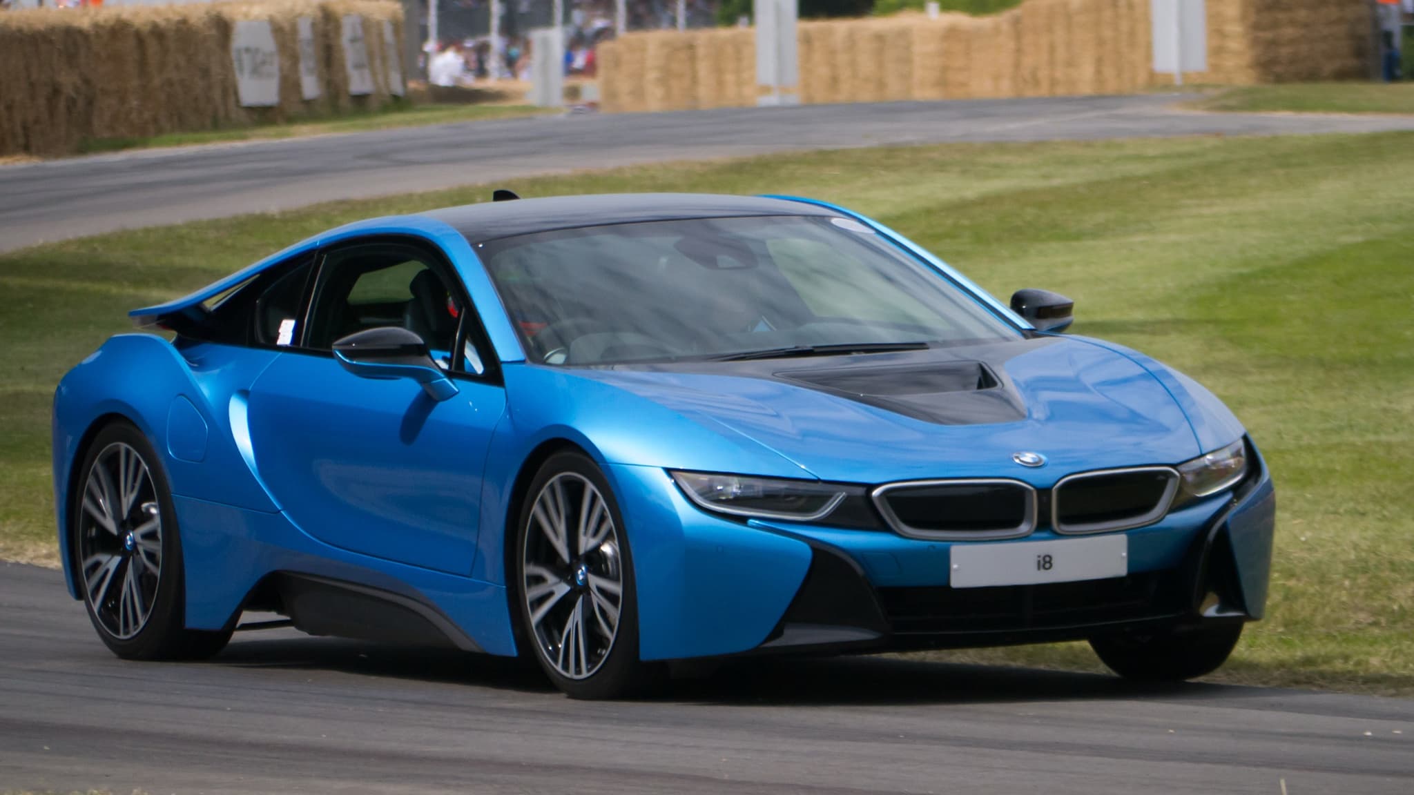 A blue BMW i8 on a hill-climb course with straw bales behind, seen from the front three-quarter.