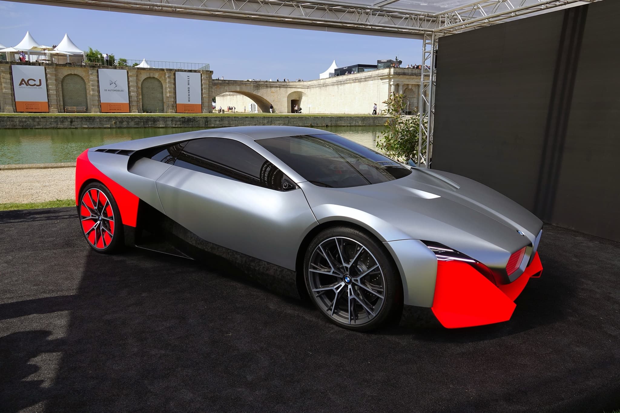The silver and red BMW Vision M NEXT concept on a black display floor under a canopy, beside a canal and a château terrace. Opens a larger view