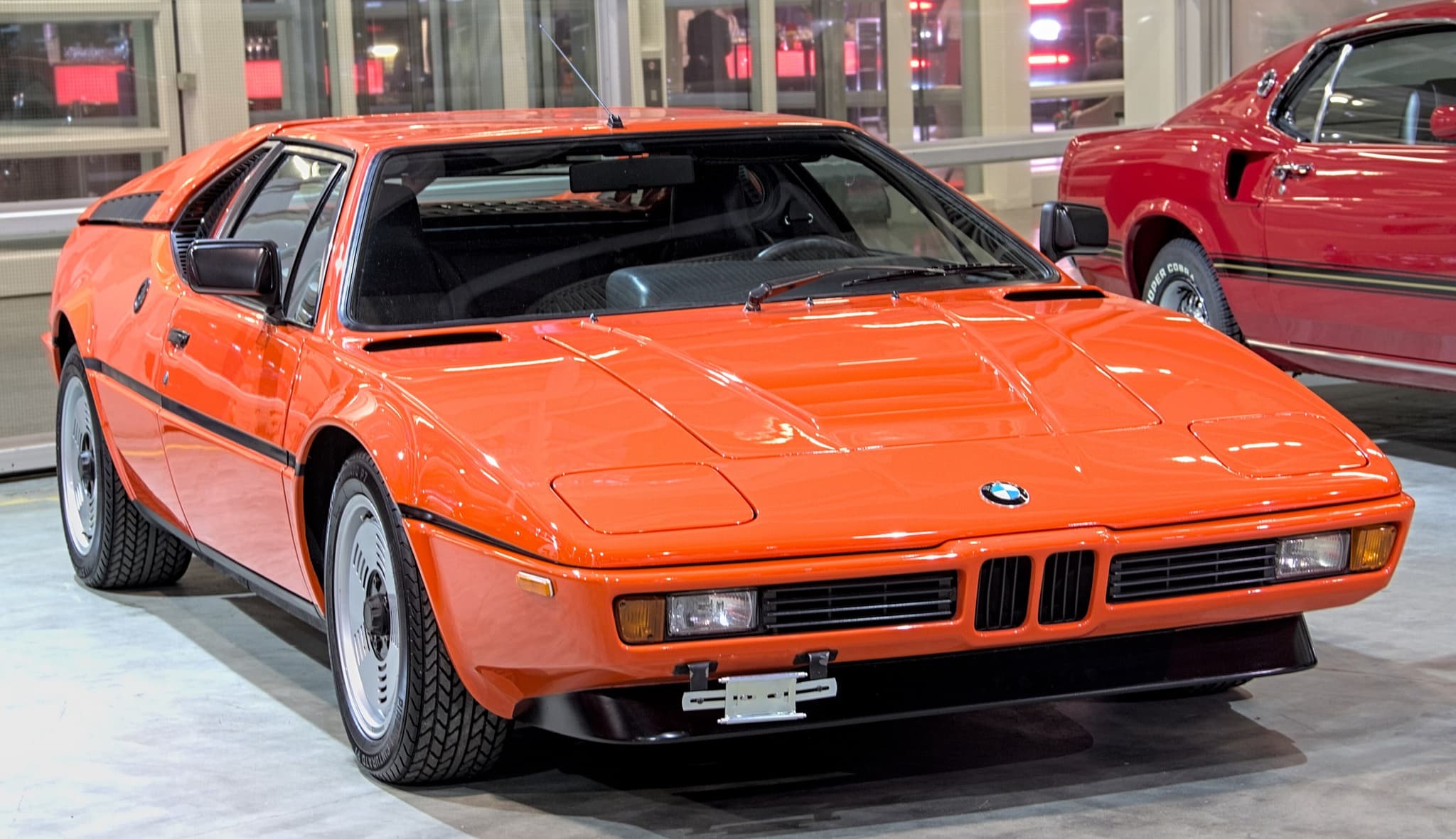An orange BMW M1 seen from the front three-quarter on a pale show floor: a low wedge nose with a small twin-kidney grille, flush headlamp covers, a black stripe along the flank and silver slotted wheels, beside a red Ford Mustang.