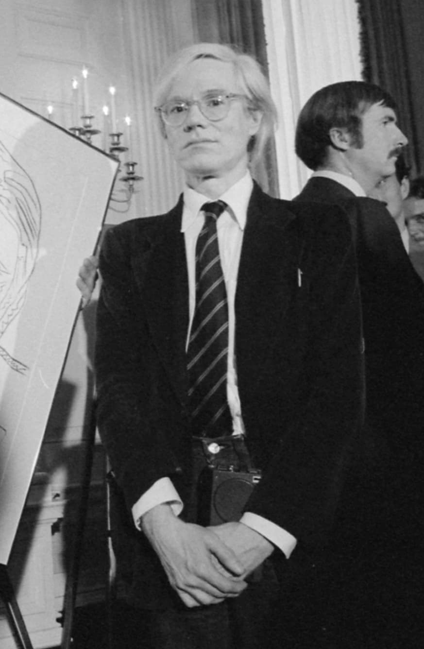 A black and white photograph of Andy Warhol in a dark suit and striped tie, hands folded, beside a framed drawing on an easel, with two men behind him.