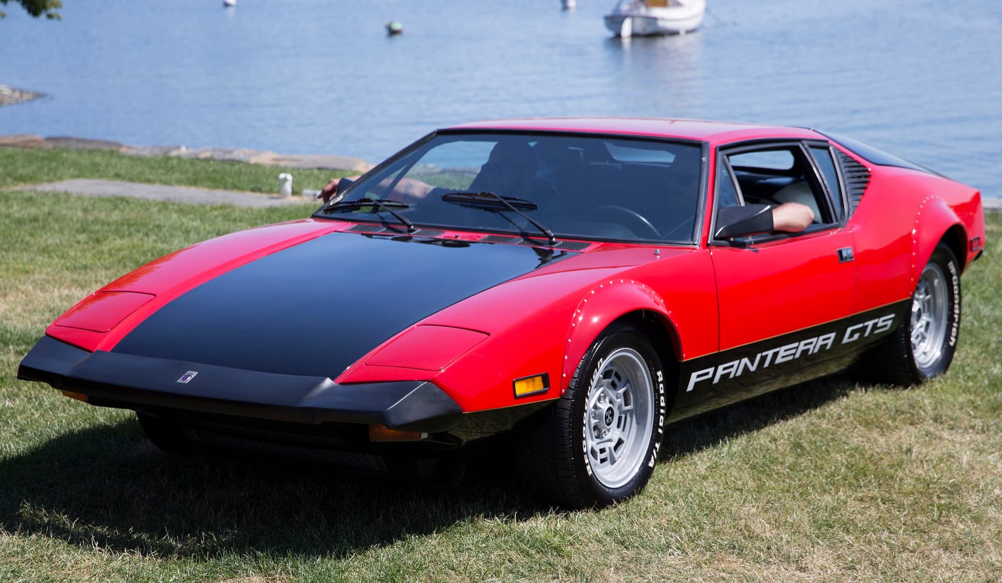 A red and black De Tomaso Pantera GTS on grass beside the water, seen from the front three-quarter.