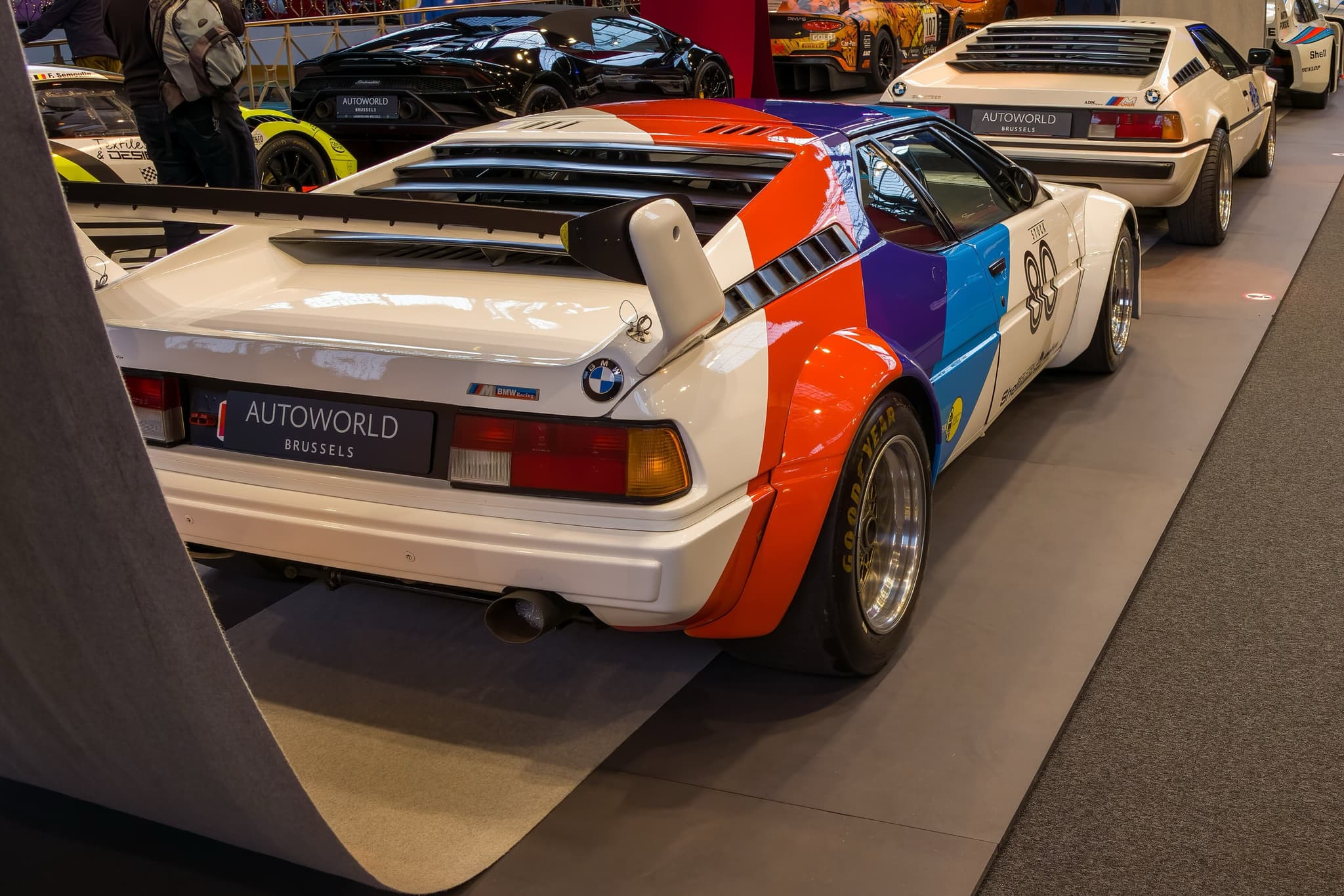 A white BMW M1 Procar in blue, violet and red stripes from the rear three-quarter in a museum, with its rear wing, flared arches and racing wheels. Opens a larger view