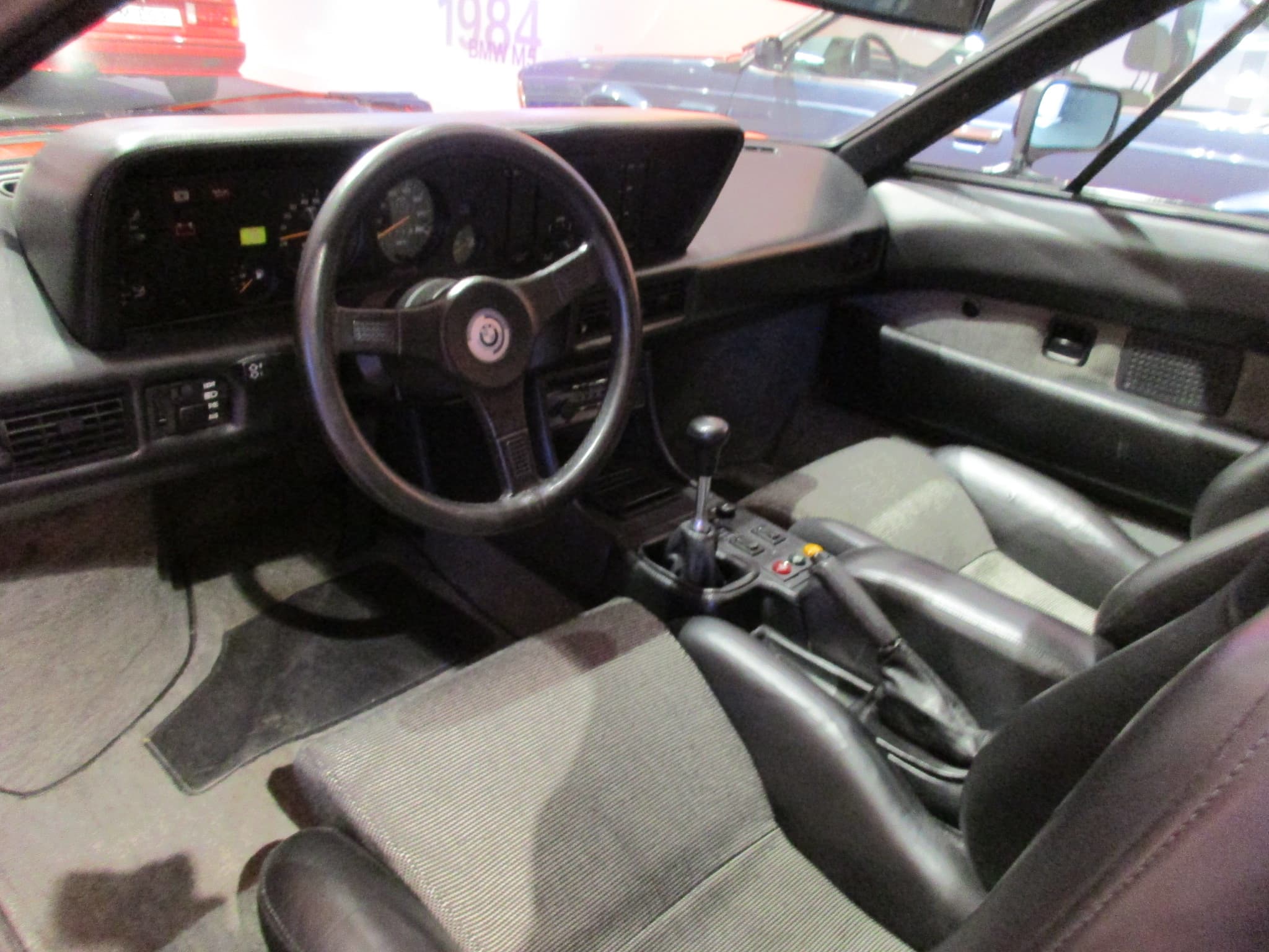 The cabin of a BMW M1 seen from the open driver's door: three-spoke steering wheel, instrument binnacle, centre console and black seats with striped cloth centres. Opens a larger view