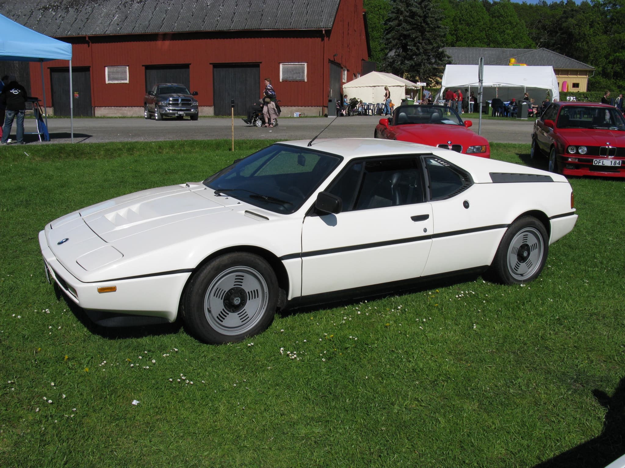 A white BMW M1 road car parked on grass at a car meeting, seen from the front three-quarter, with a barn and tents behind. Opens a larger view