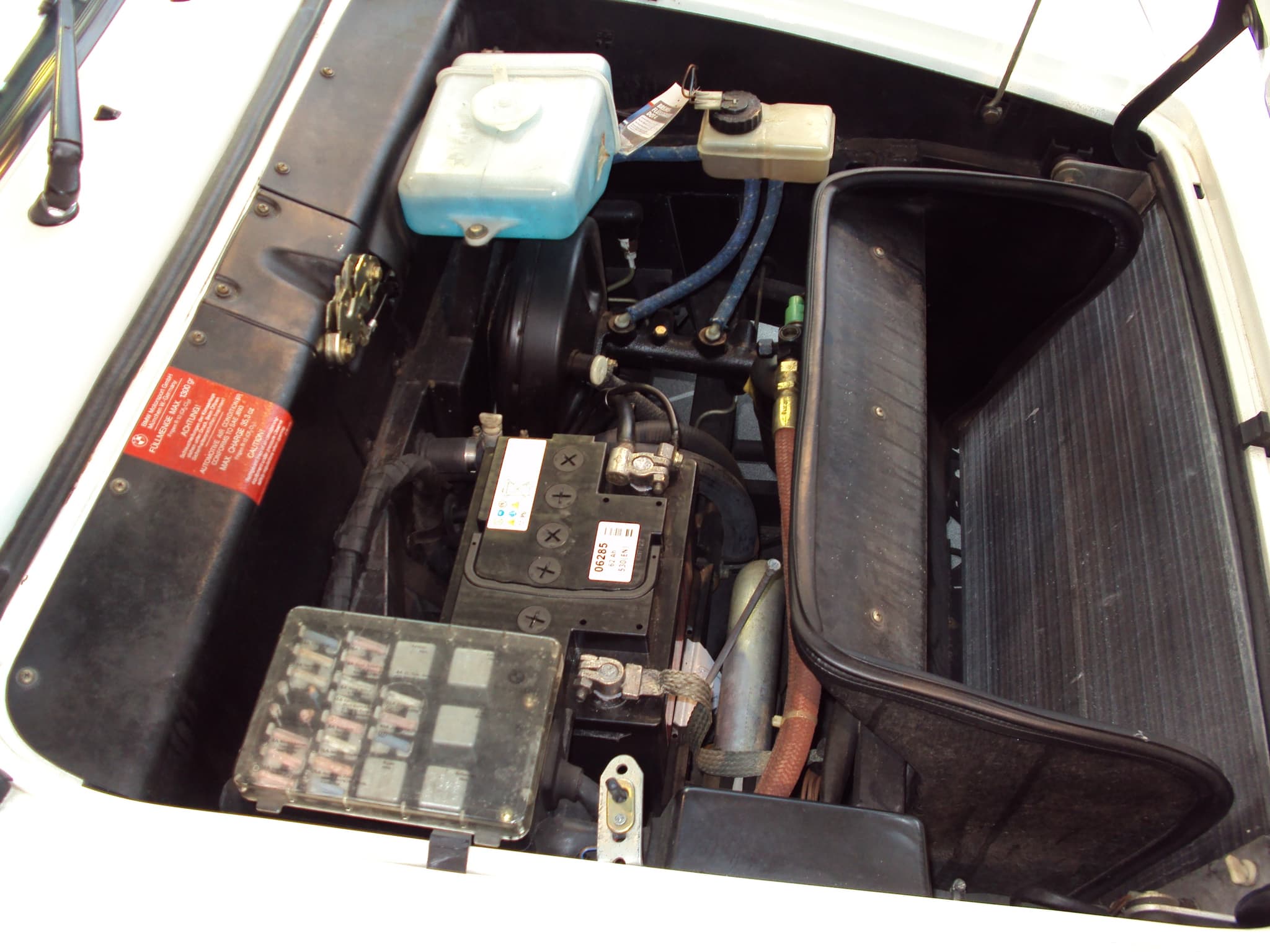 The front compartment of a white BMW M1 with the bonnet open: fluid reservoirs, battery, fuse box and a lined luggage tray. Opens a larger view