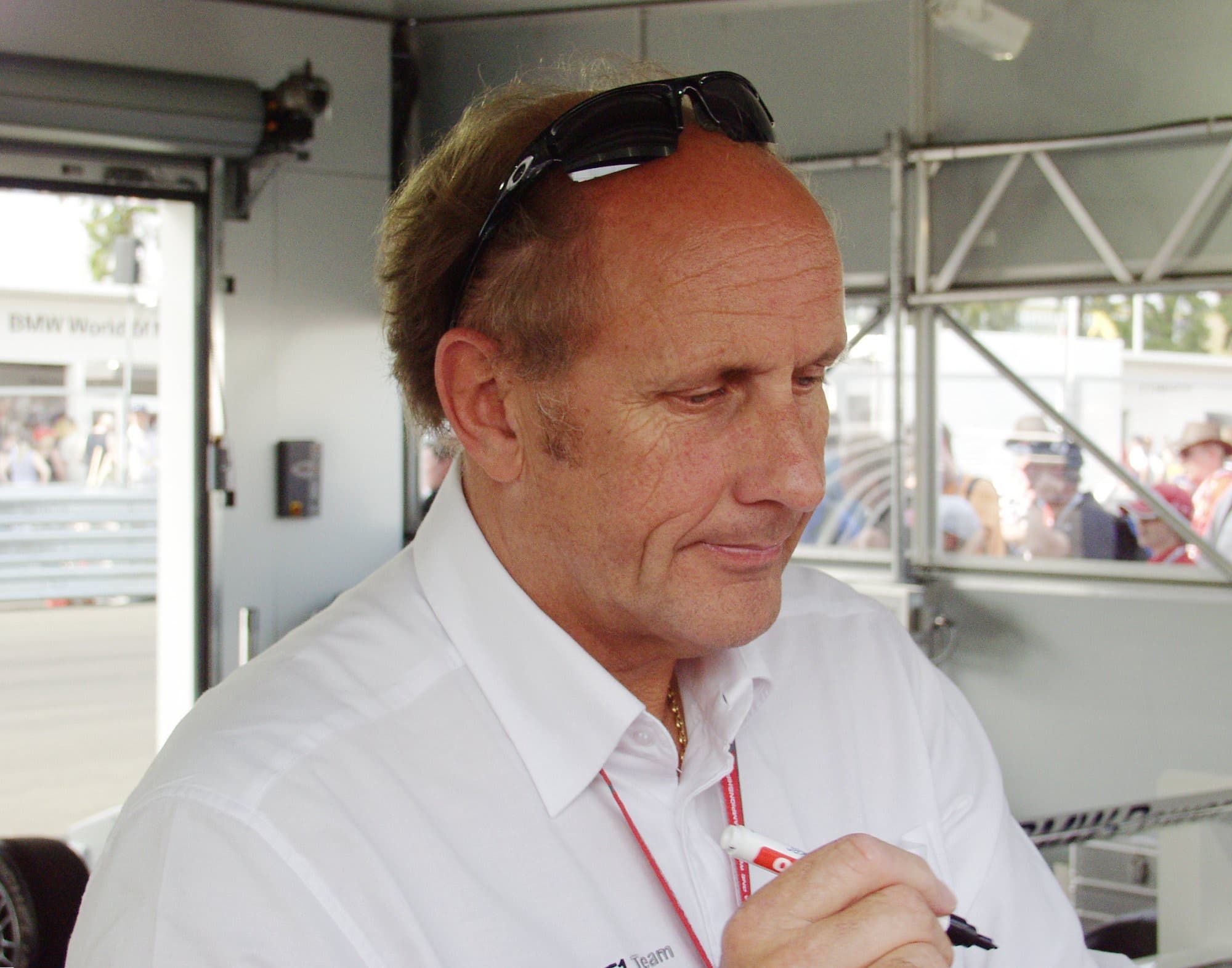 Hans-Joachim Stuck in a white shirt with sunglasses on his head, signing with a marker inside a paddock booth.