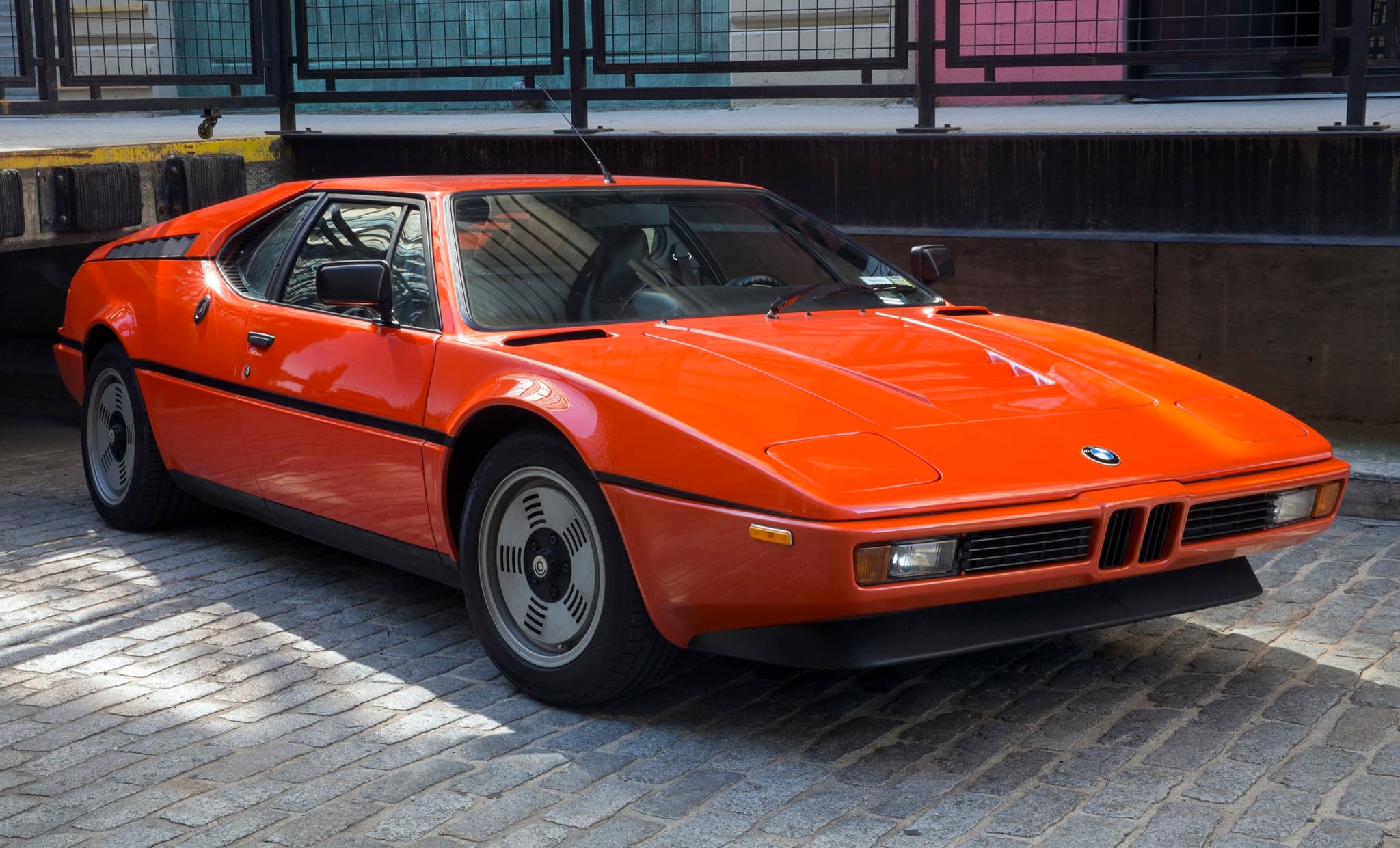 An orange BMW M1 road car parked on cobbles in low sunlight, front three-quarter from the right.