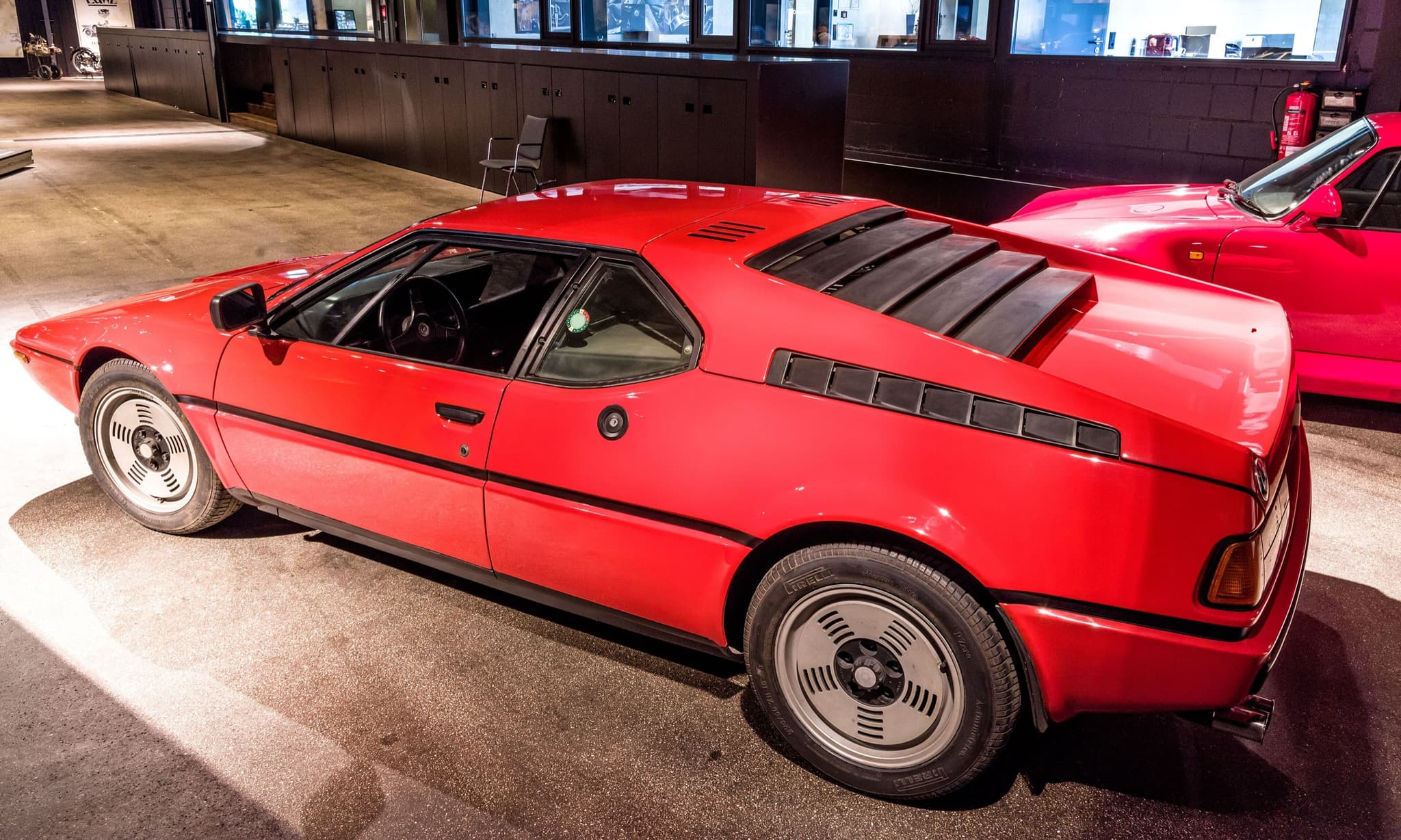 A red BMW M1 road car seen from the left and slightly behind, with the louvred rear deck, buttress louvres and the fuel filler flap.