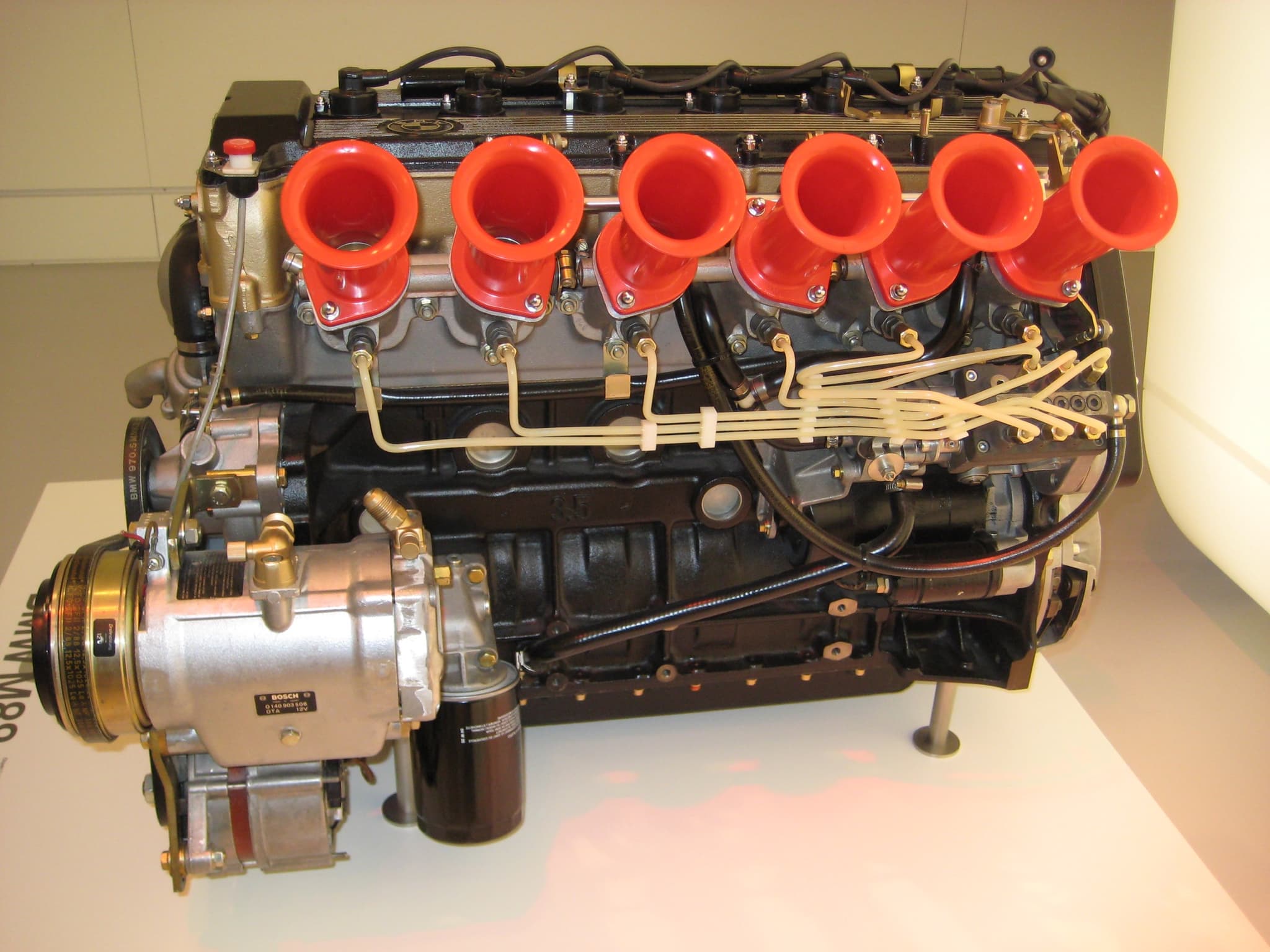A BMW M88 straight-six engine on a museum plinth, with six red intake trumpets in a row and its fuel-injection lines.