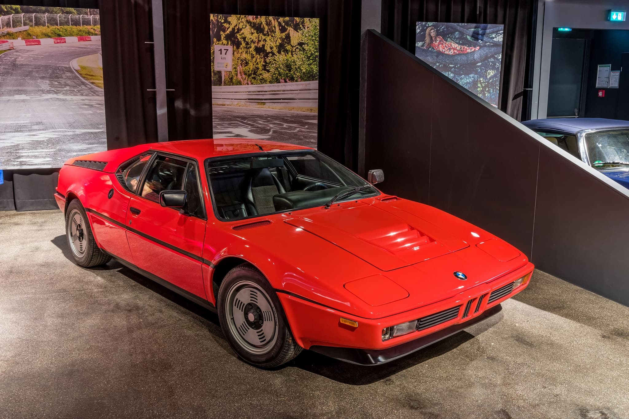 A red BMW M1 road car in a museum bay, front three-quarter, with Nürburgring photographs behind it. Opens a larger view