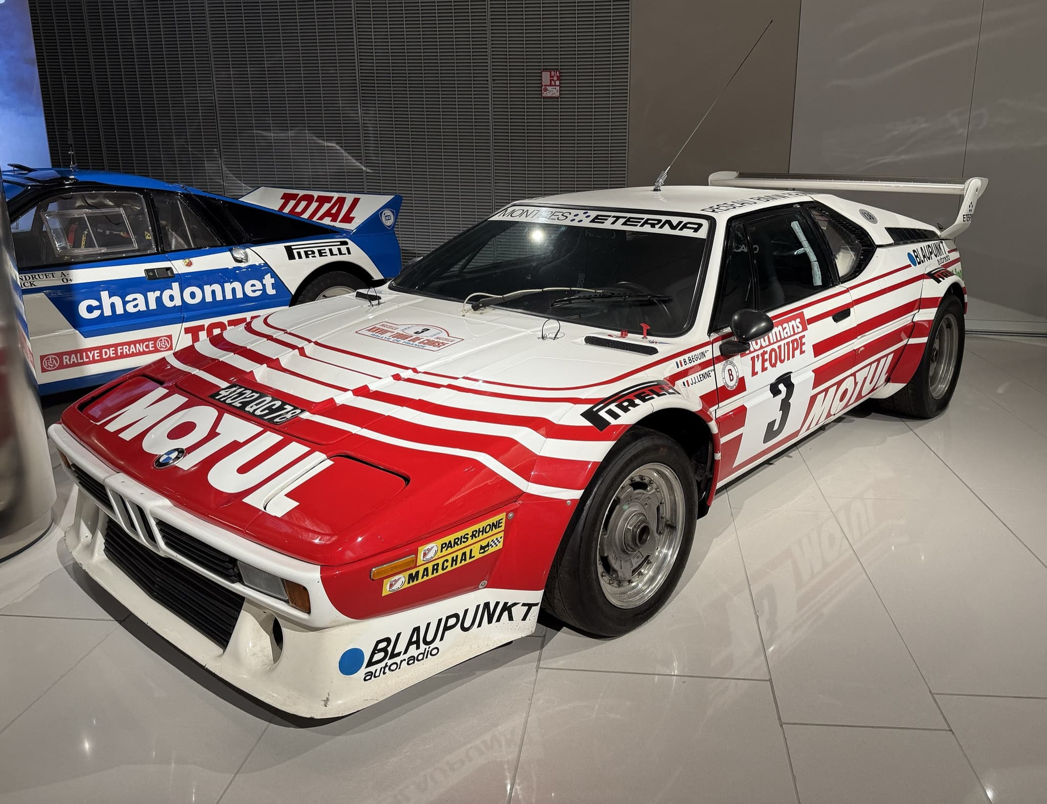 The red and white Motul rally BMW M1, number 3, in a museum, beside a blue and white rally car.