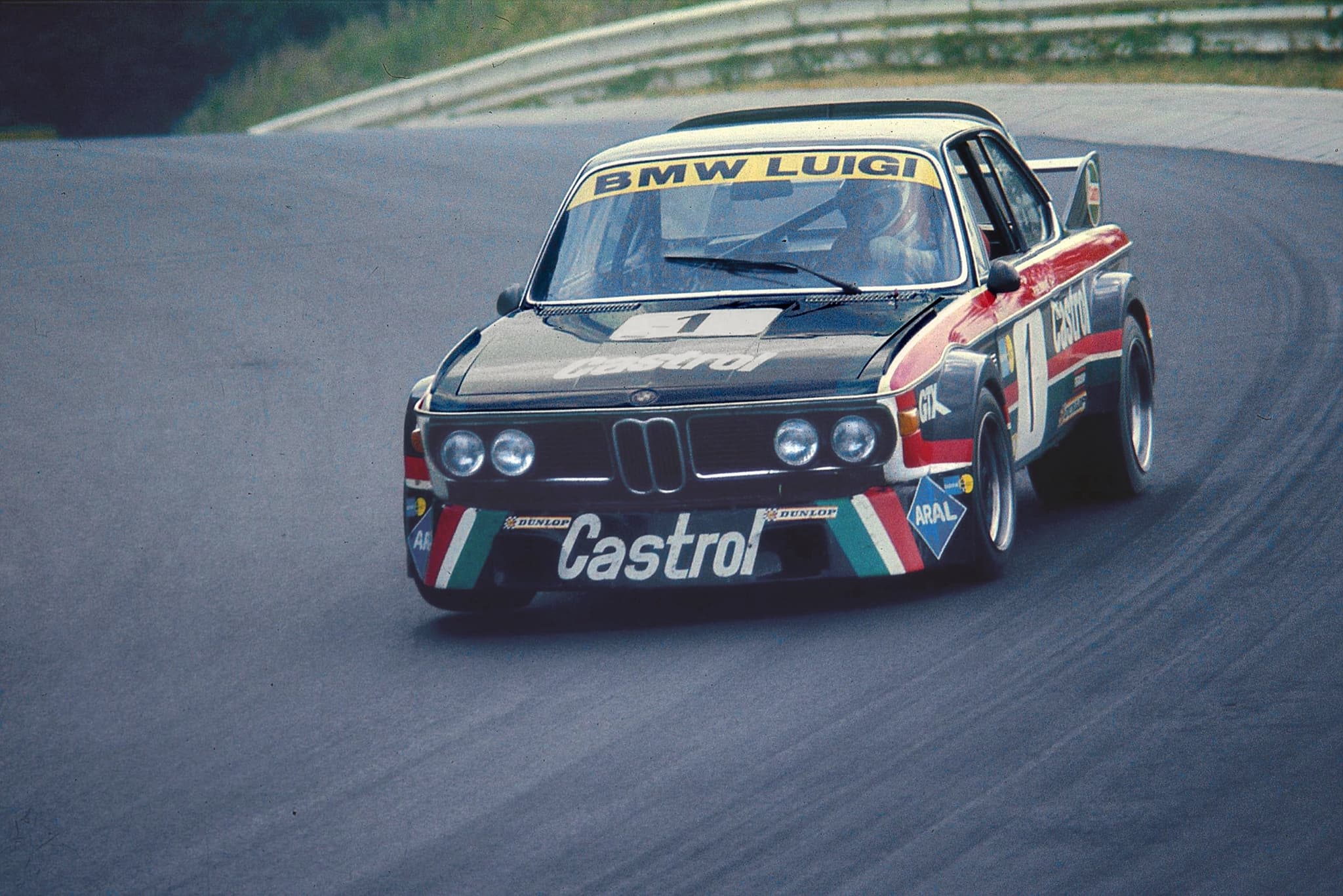 A dark BMW CSL racing car in Castrol colours with BMW LUIGI on the windscreen strip, number 1, cornering on the Nürburgring.