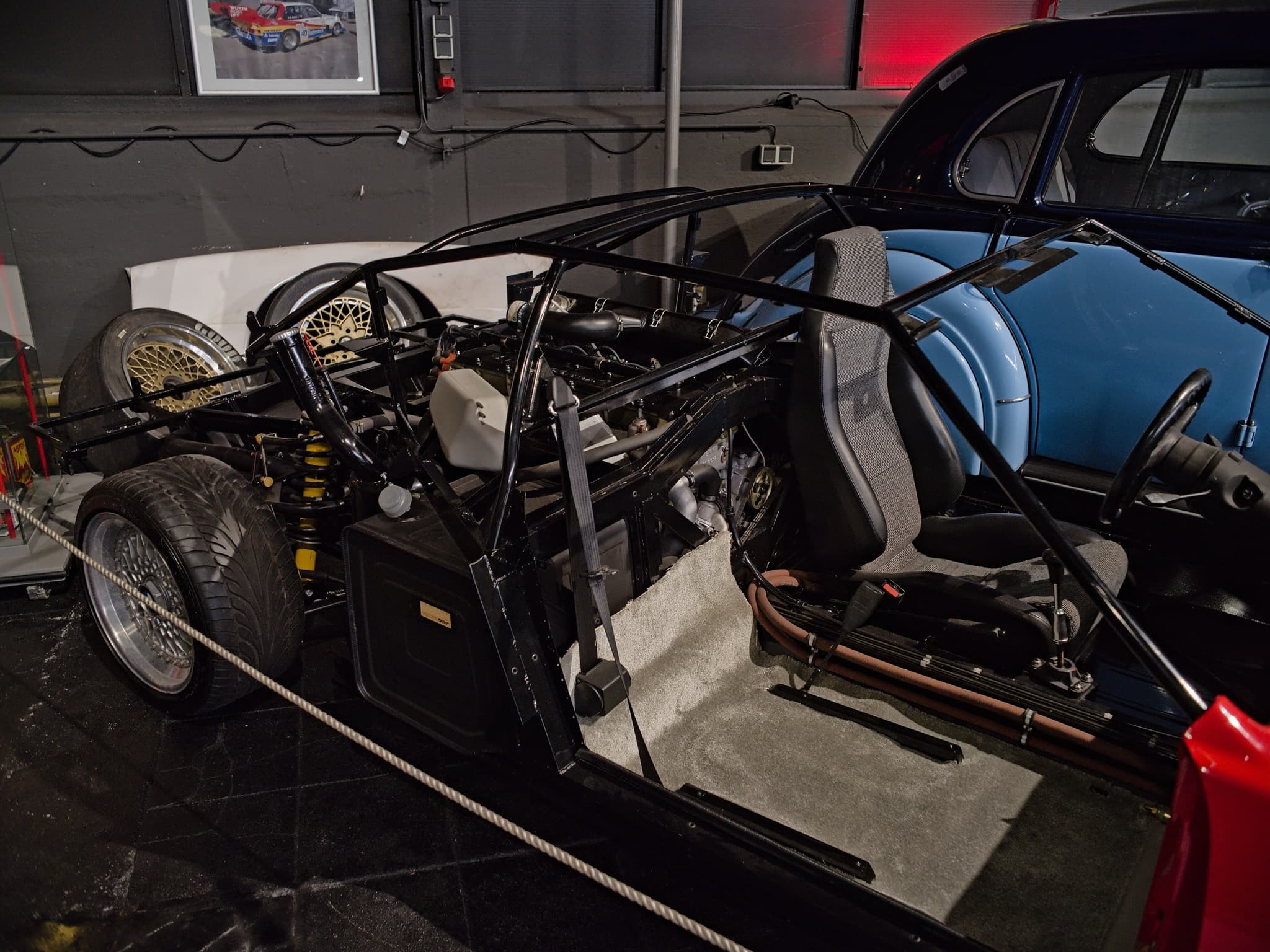 The same bodiless BMW M1 display from the rear quarter: the tubular frame with a roll cage, a racing-style seat, belts and steering wheel.