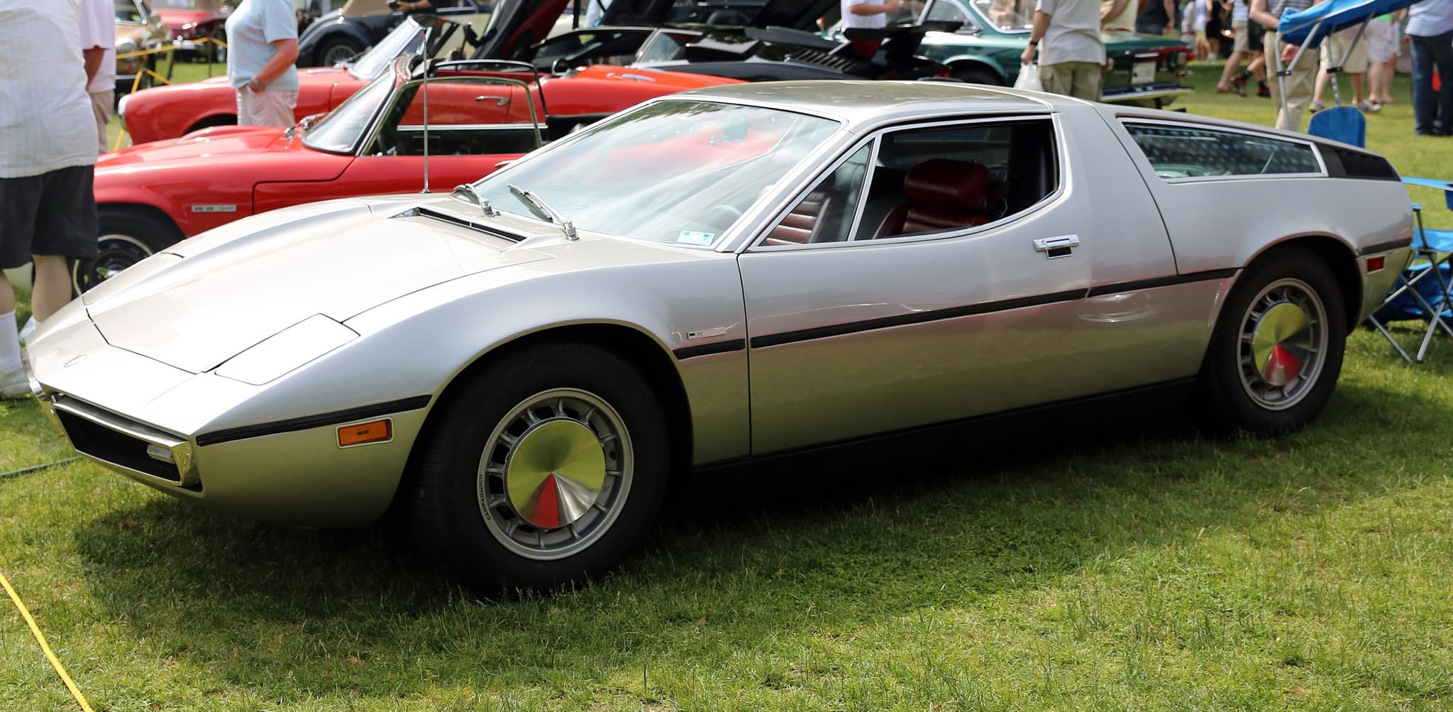 A silver Maserati Bora on grass at a concours, seen from the front three-quarter, with other cars behind.