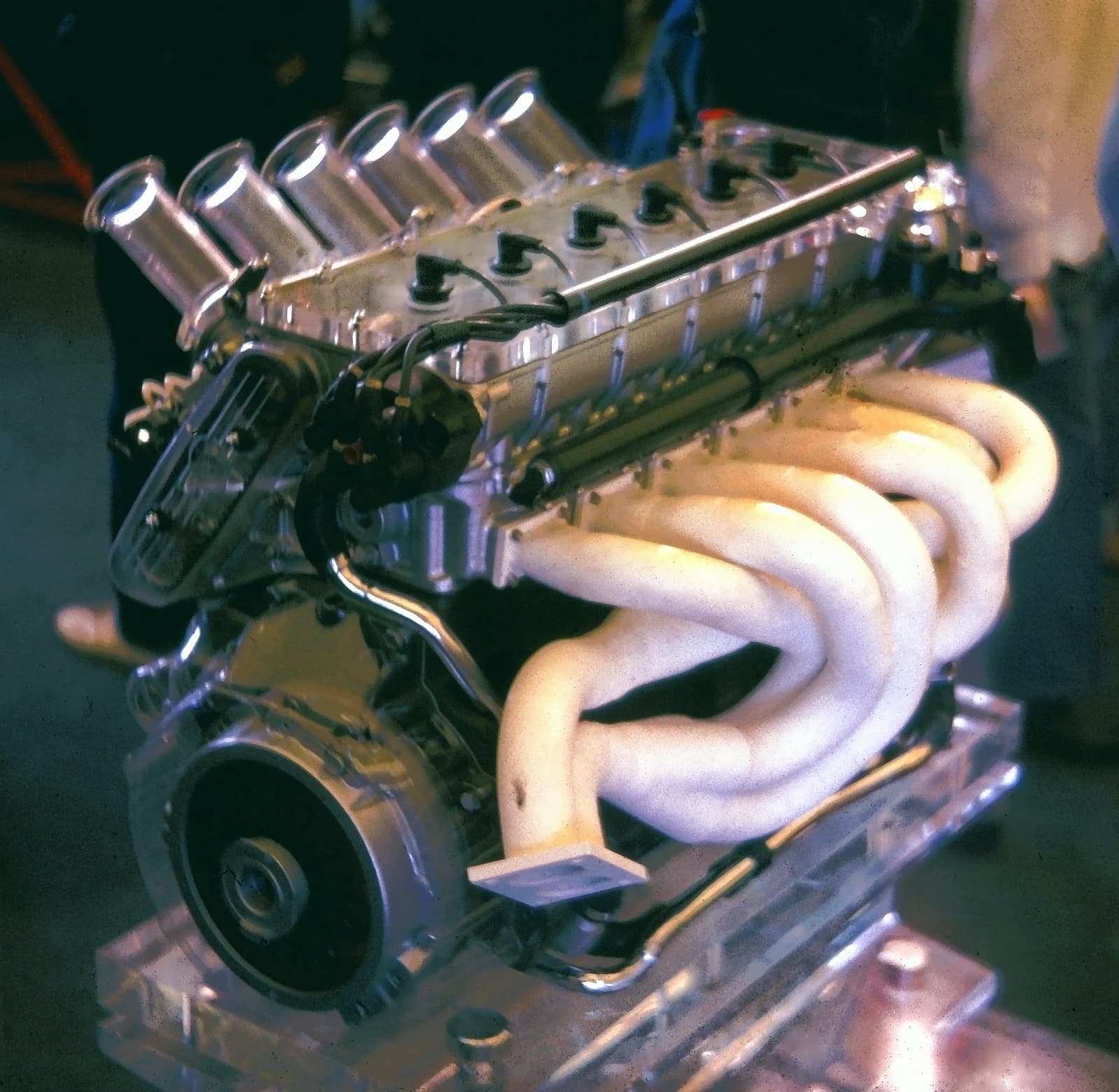 A BMW M1 engine on a display stand, partly cut away in clear Perspex, with polished intake trumpets and a wrapped exhaust manifold.