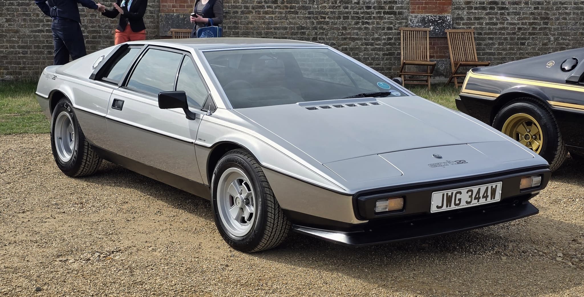 A silver Lotus Esprit S2.2 on gravel in front of a brick wall, seen from the front three-quarter, with a black and gold Esprit beside it.