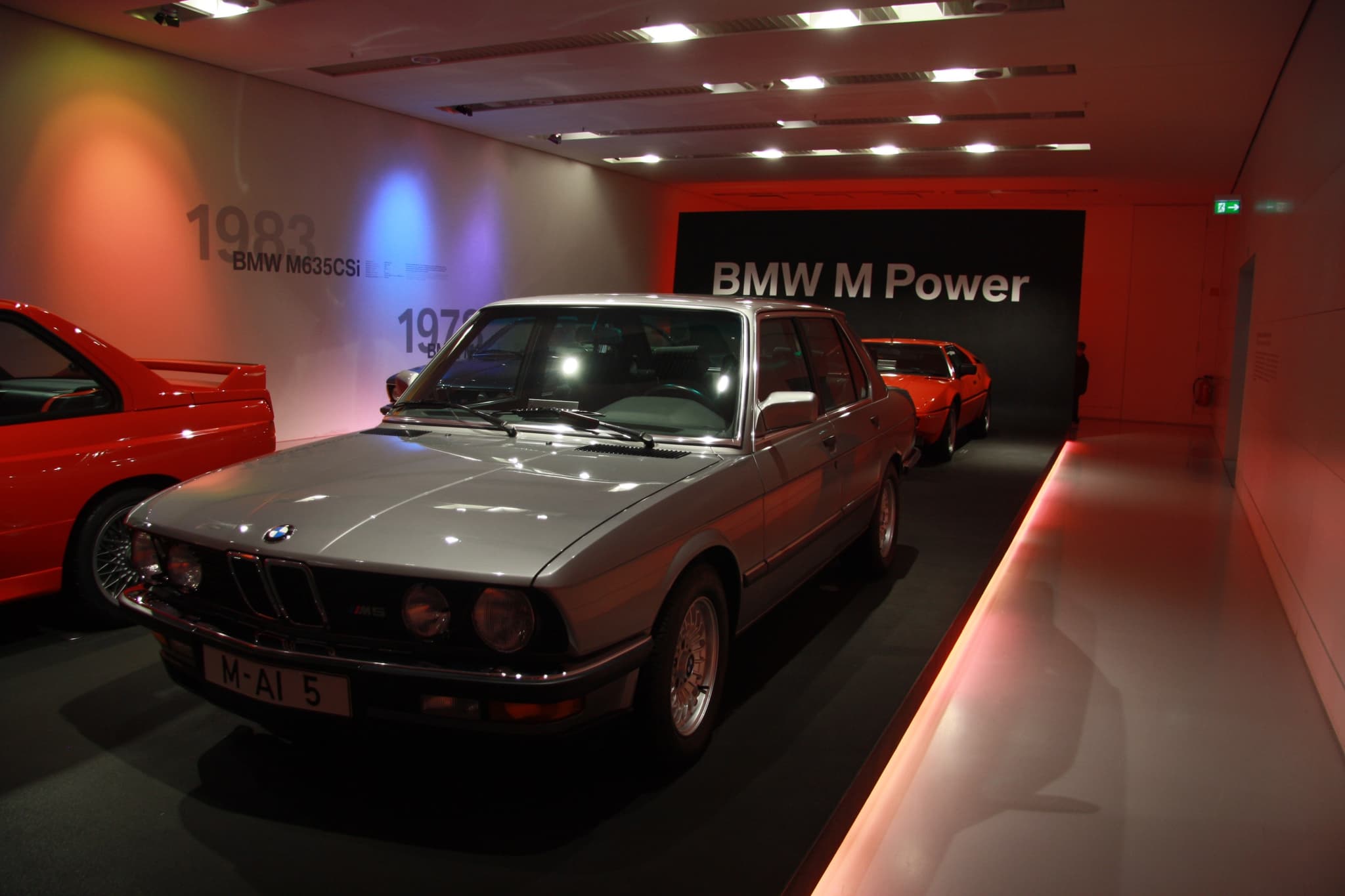 A silver BMW M5 (E28) in a museum room, with an orange BMW M1 and a red M3 beyond.