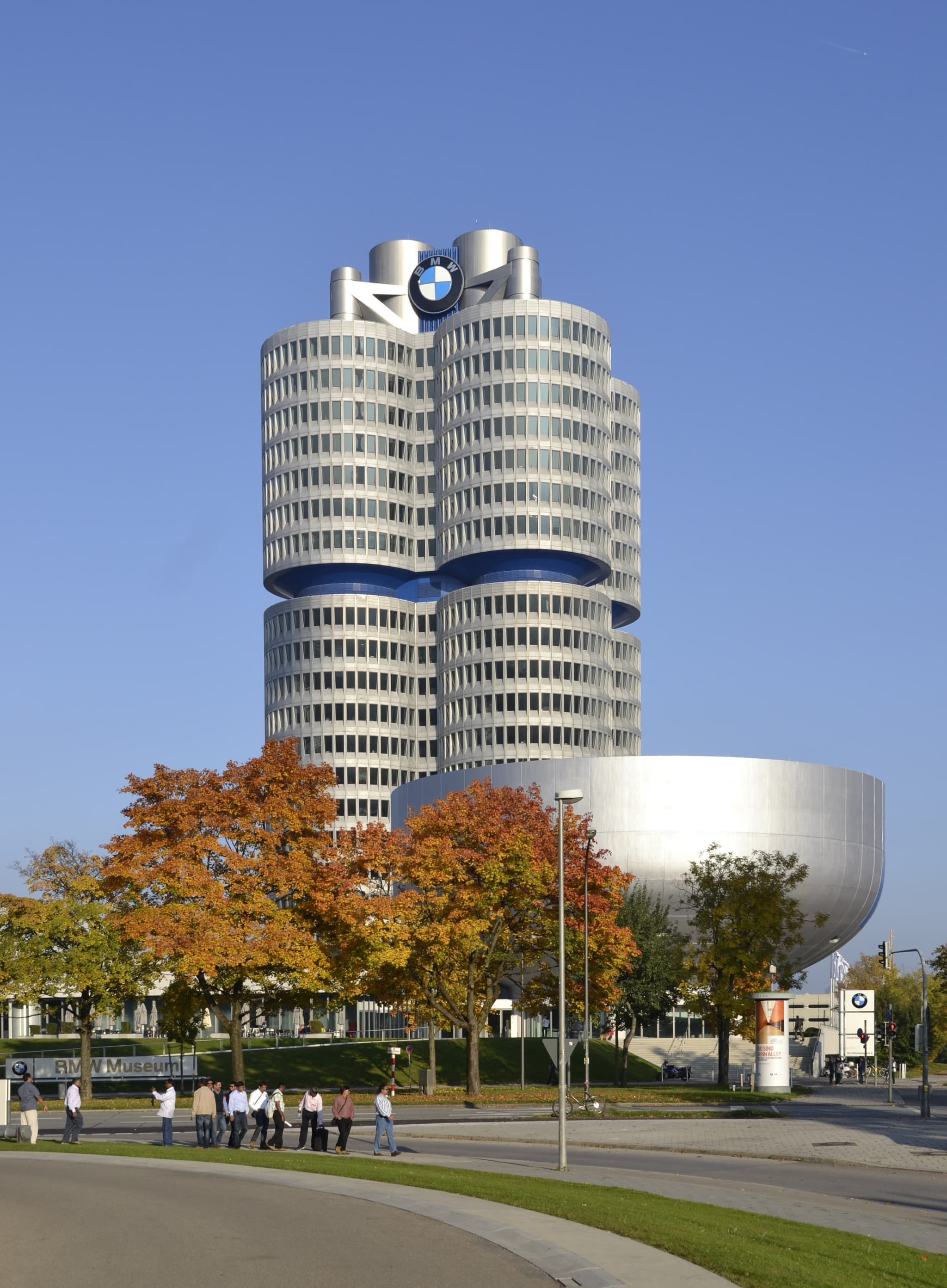 A tall office tower of four joined silver cylinders with a BMW roundel on its roof, beside a low silver bowl-shaped building, behind trees in autumn colours.