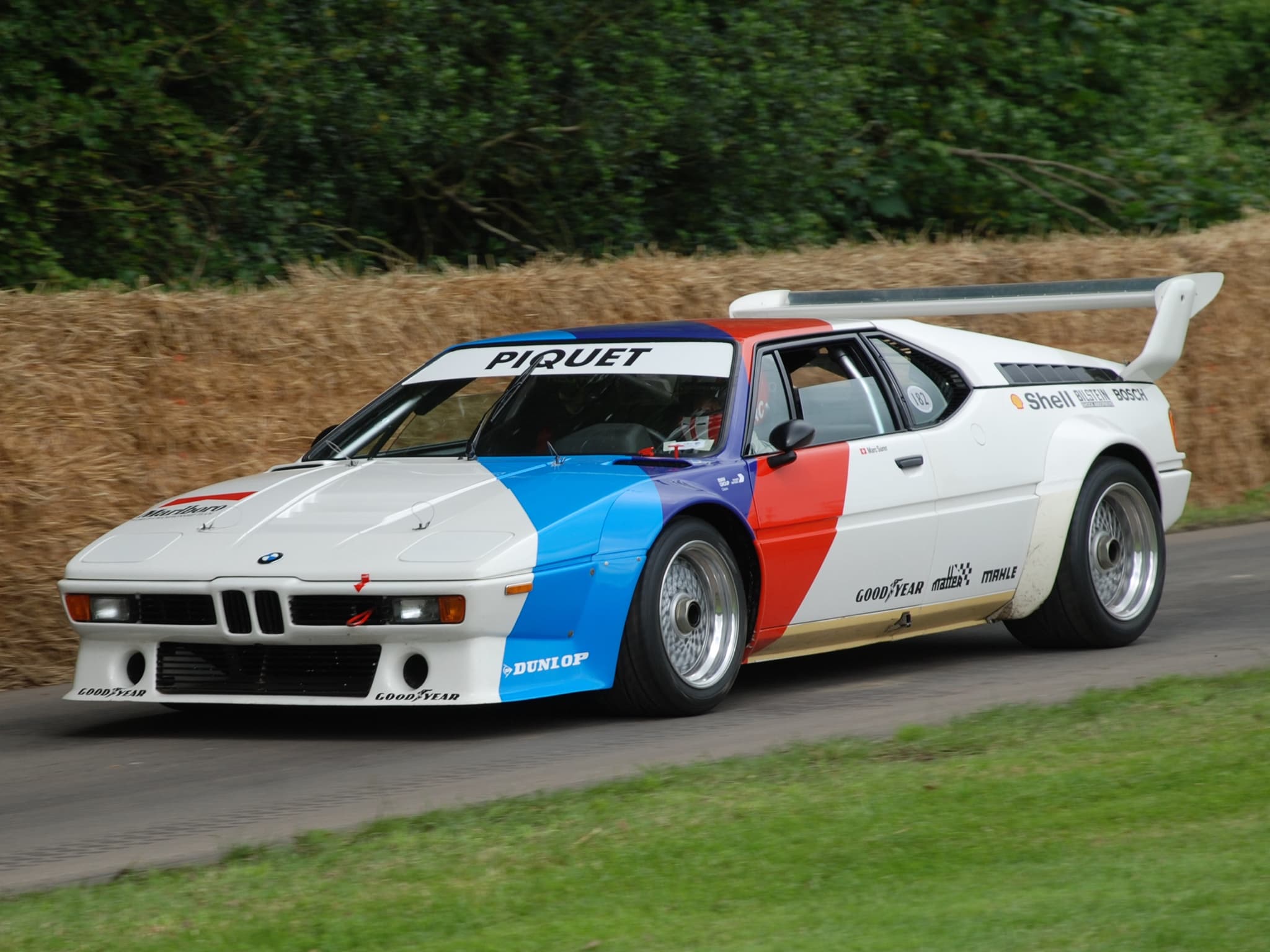 A white BMW M1 Procar with blue, violet and red stripes and PIQUET on the windscreen strip, on the Goodwood hill course beside straw bales.