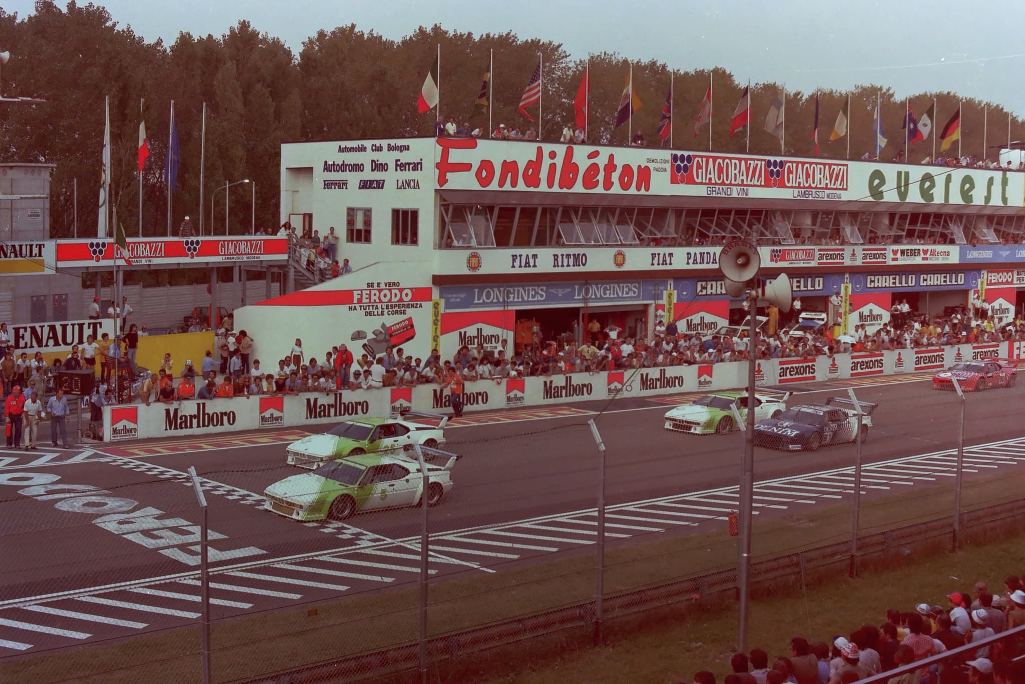Procars on the start and finish straight at Imola in 1980, seen through a catch fence, with the pit building's advertising and spectators. Opens a larger view