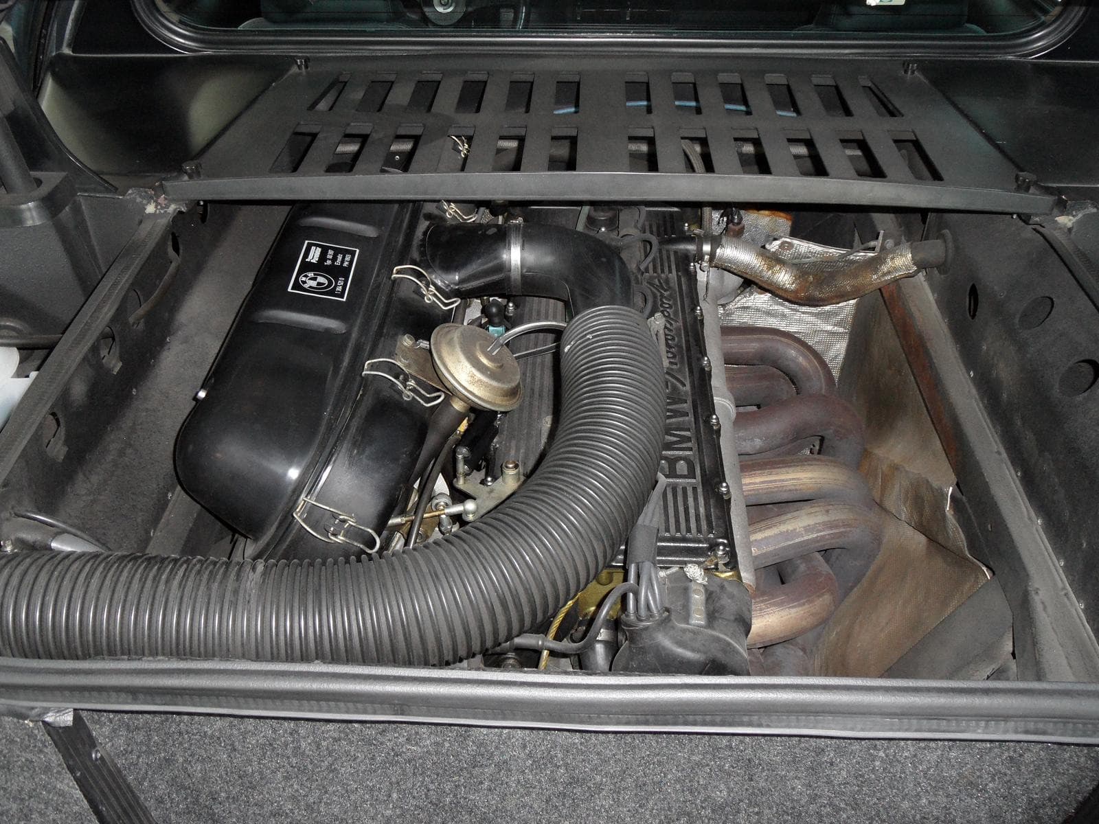 The engine bay of a BMW M1 seen from behind, with the cam cover lettered BMW Motorsport, a black filter box and a corrugated air hose.