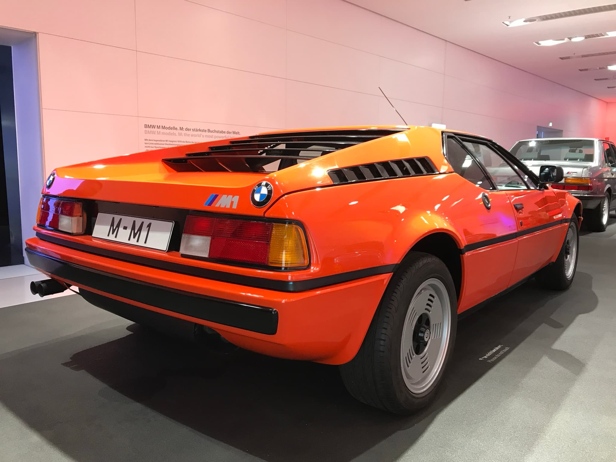 An orange BMW M1 road car in a museum hall, seen from behind and to the right, with slats over the engine cover and louvres along its flank.