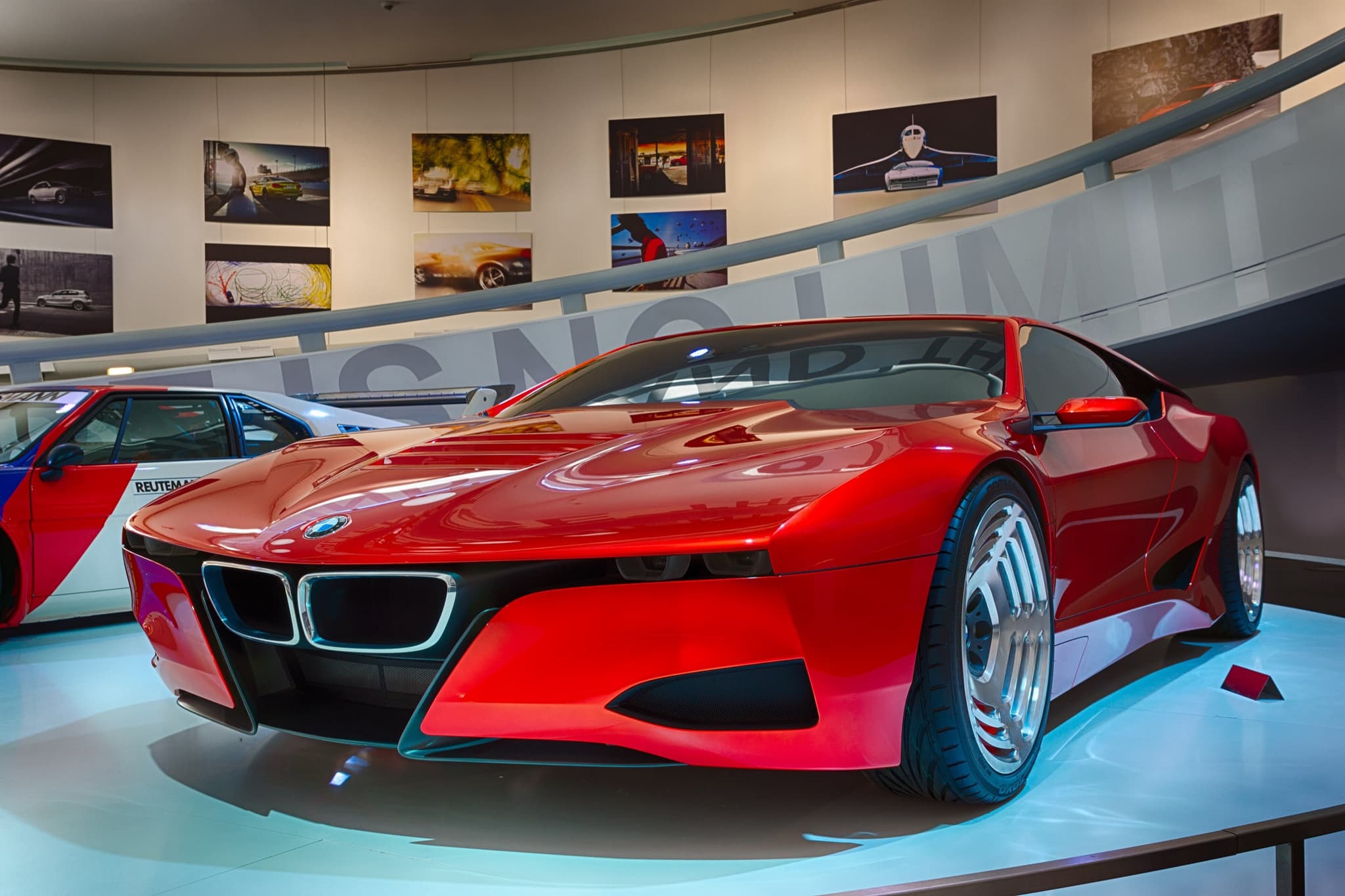 The red BMW M1 Hommage concept on a lit museum ramp, front three-quarter, with a racing M1 at the left.