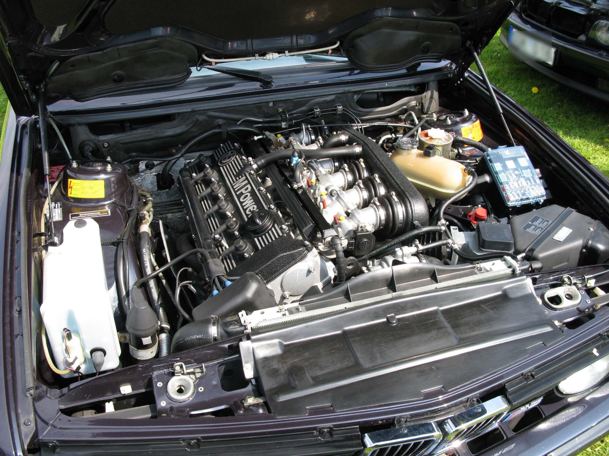 The engine bay of a BMW M5 (E28) with an M Power cam cover over its six individual throttle bodies.