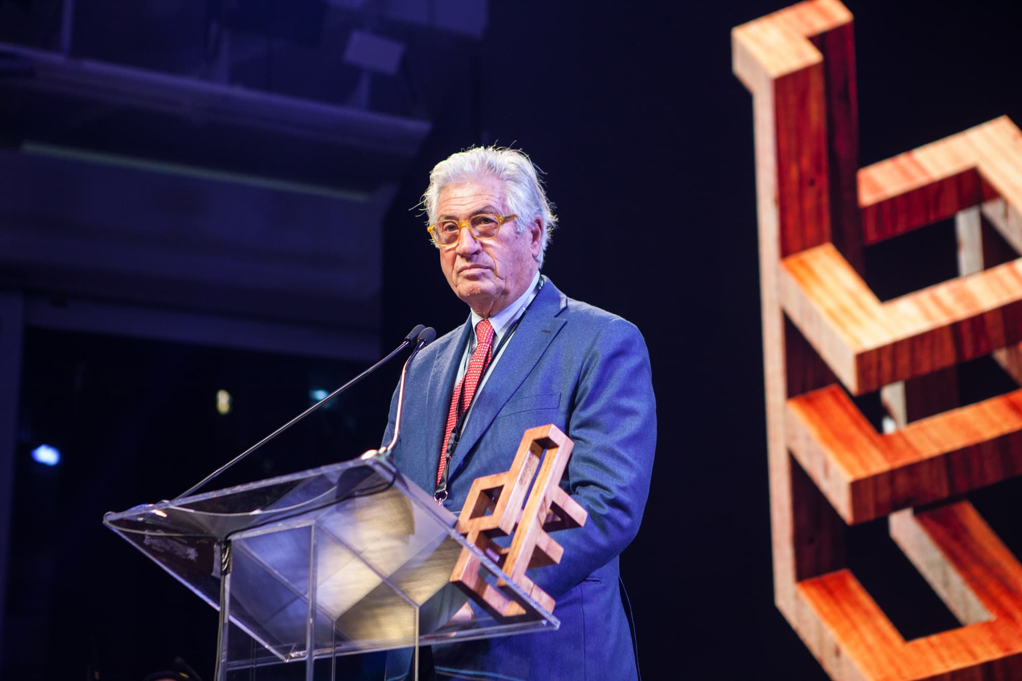 A grey-haired man in glasses, a blue suit and a red tie, standing at a clear lectern on a dark stage with a wooden award in front of him.
