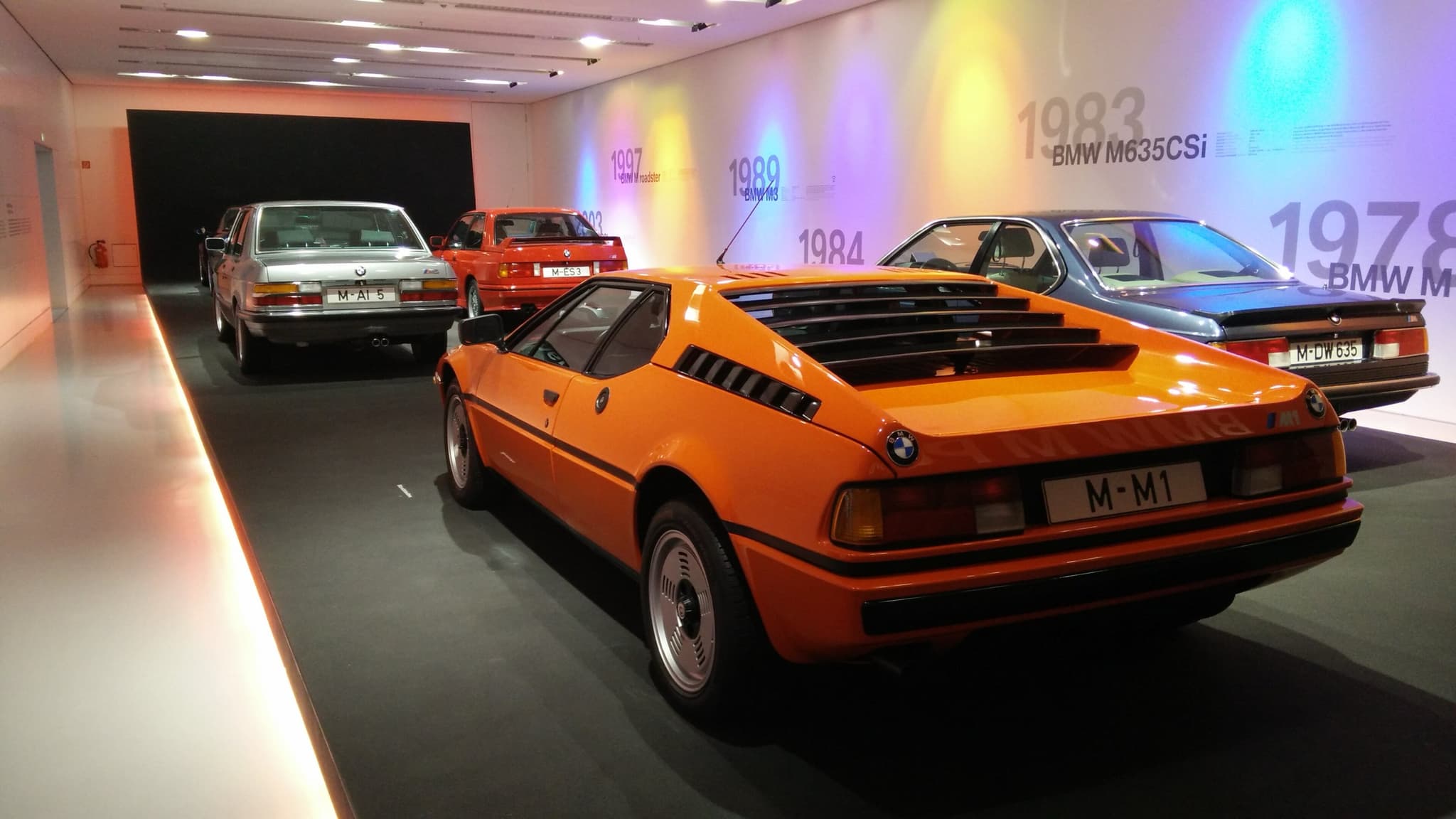 An orange BMW M1 seen from behind in a museum room, beside an M635CSi with an M5 and an M3 beyond and years printed on the wall.
