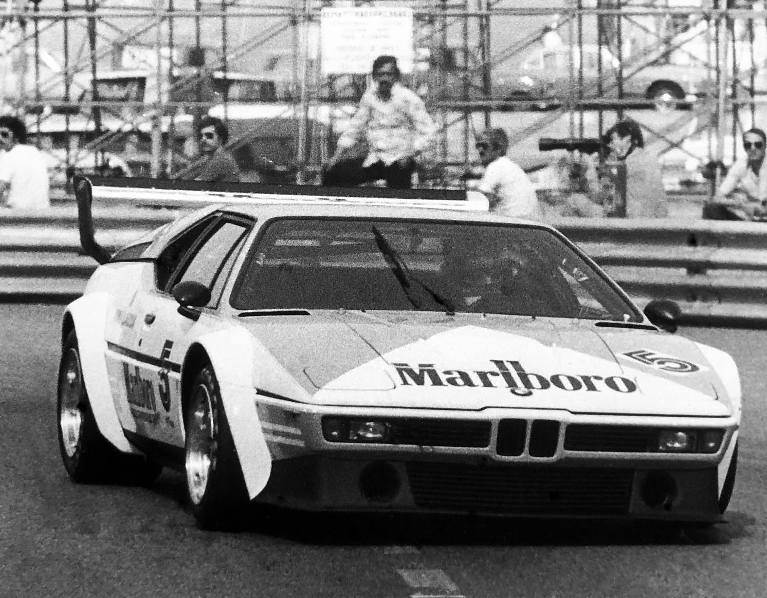 A black and white photograph of a Procar in Marlboro colours, number 5, coming through a Monaco corner, photographers behind the barrier.