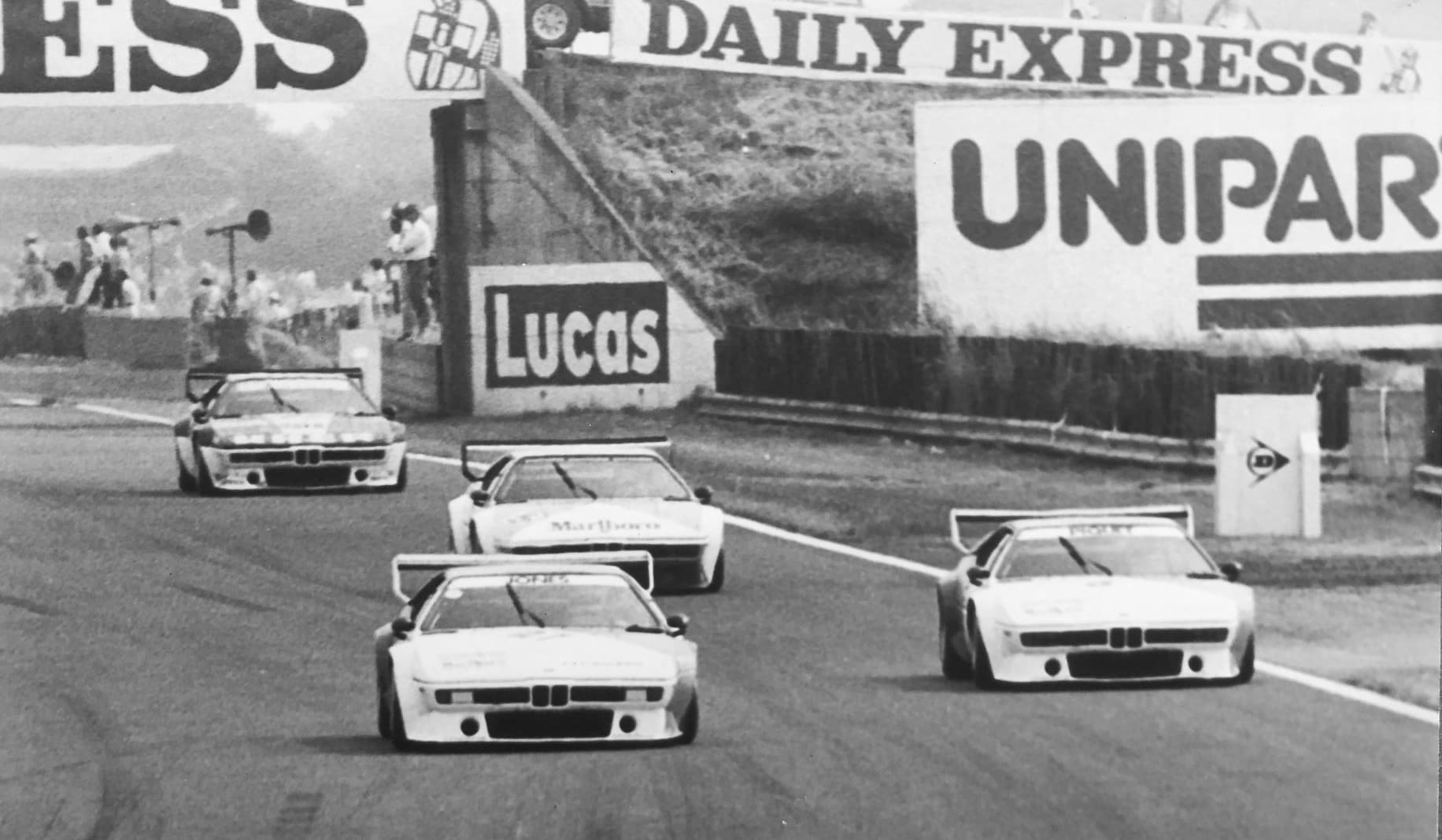 A black and white photograph of four Procars racing at Silverstone, with period trackside advertising behind.