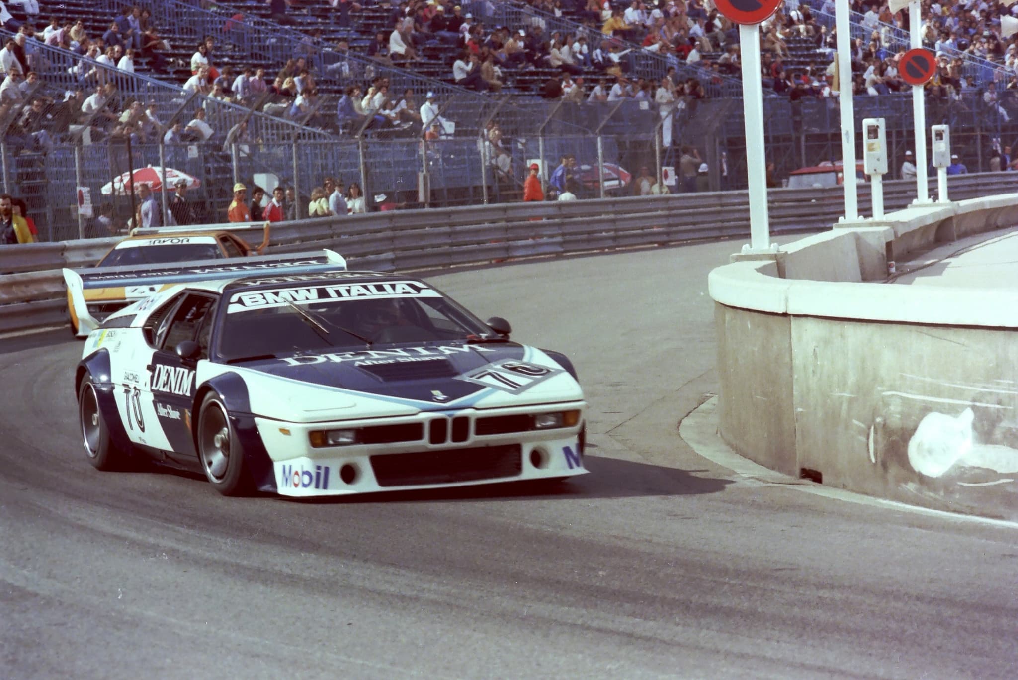 A white and dark blue Procar lettered DENIM and BMW ITALIA, number 70, cornering at Monaco in front of a full grandstand, with a second Procar behind.