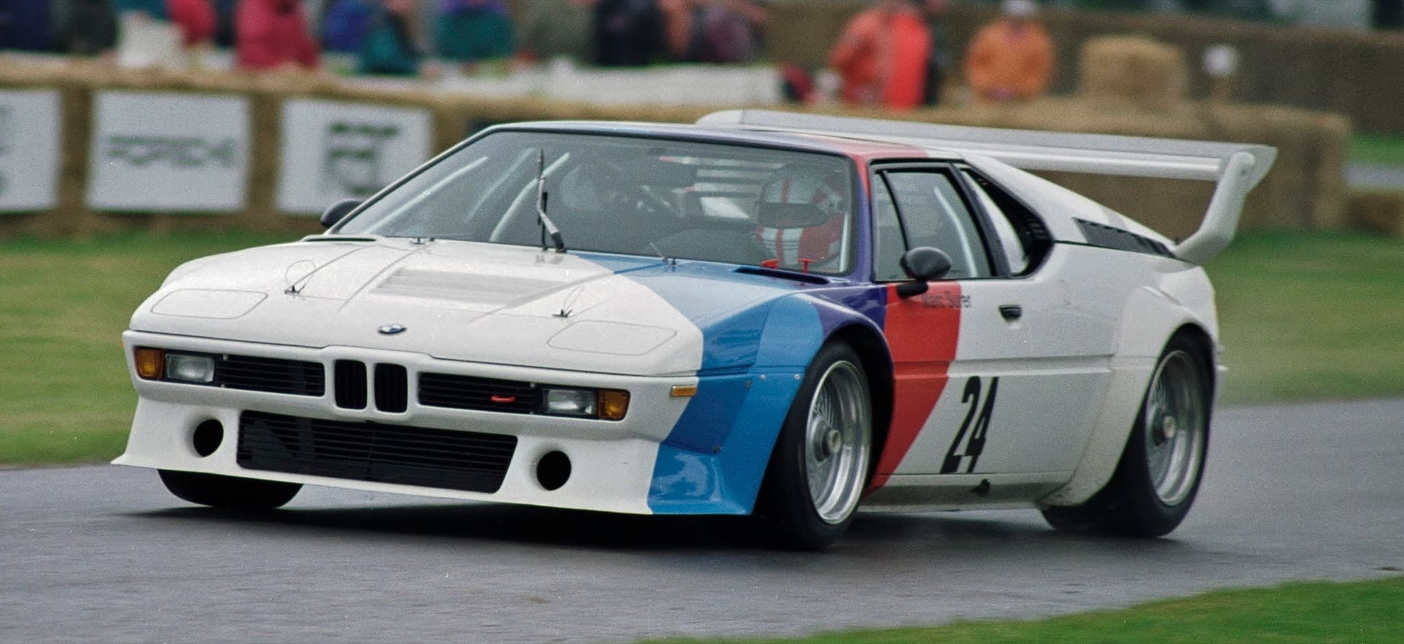 A white BMW M1 Procar with blue, violet and red stripes and the number 24, cornering on a wet course with blurred spectators behind.