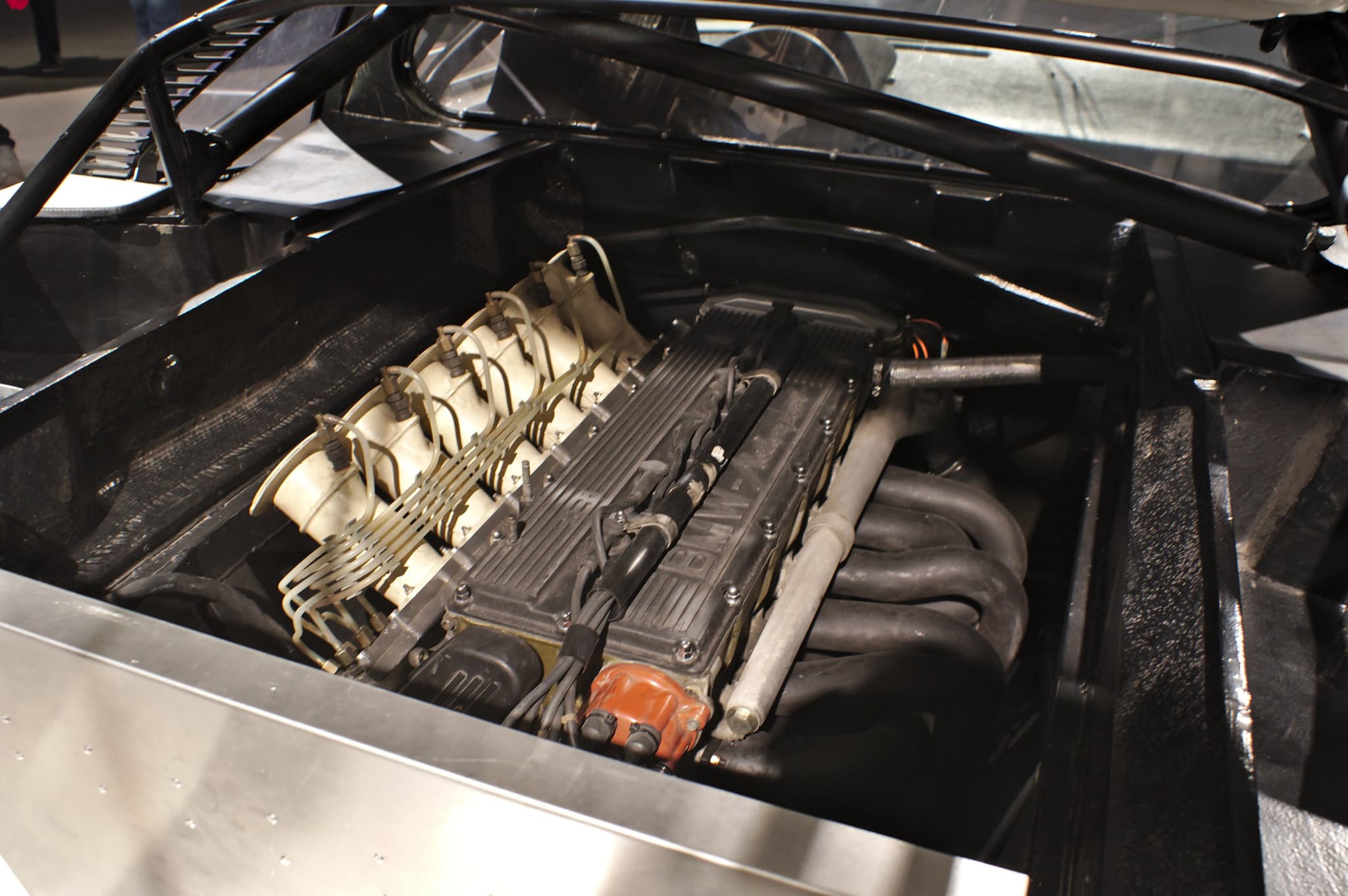 Looking down into a racing BMW M1's engine bay: the six-cylinder with its injection system inside a roll cage and bare aluminium panels.