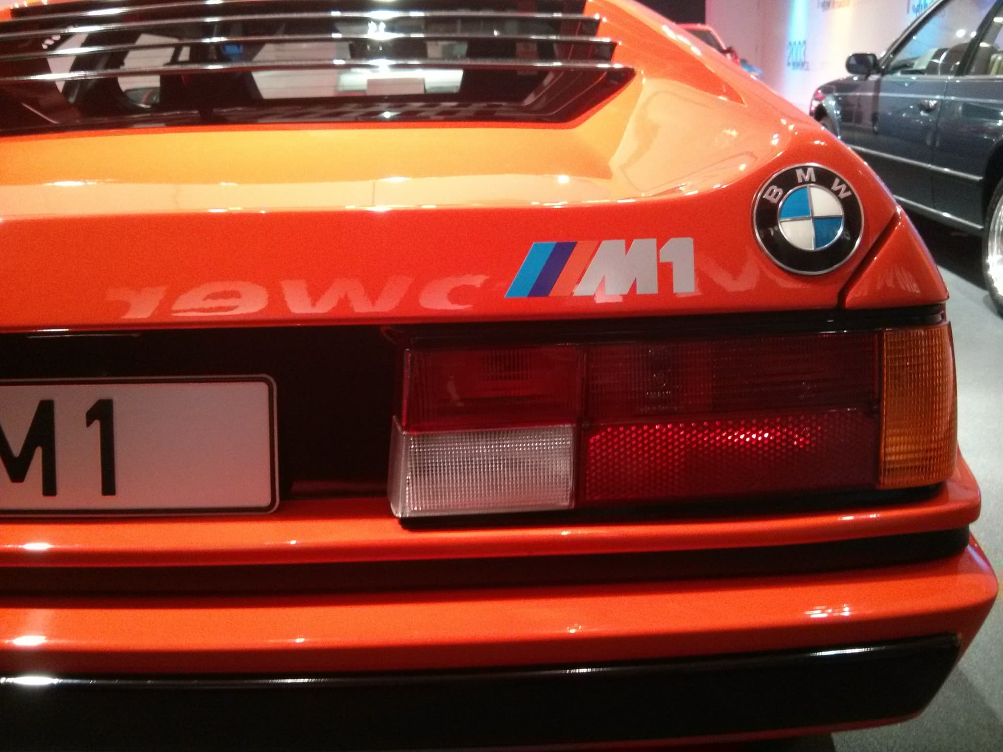 Close-up of an orange BMW M1's tail: the M1 badge with the M stripes, a corner roundel and a tail lamp. Opens a larger view