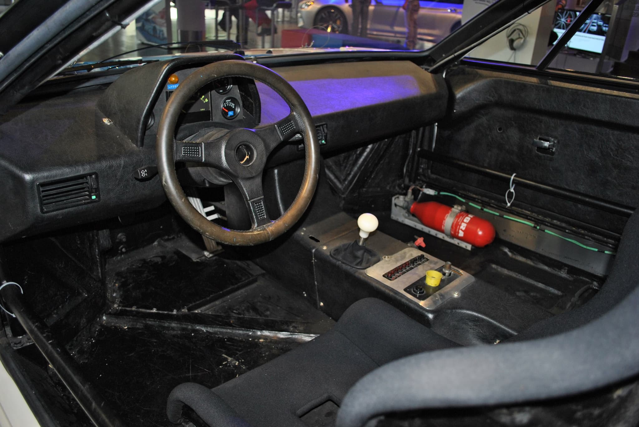 The cockpit of a BMW M1 Procar: a three-spoke wheel, racing gauges, a white gear knob, a switch panel, a fire extinguisher and a racing seat. Opens a larger view