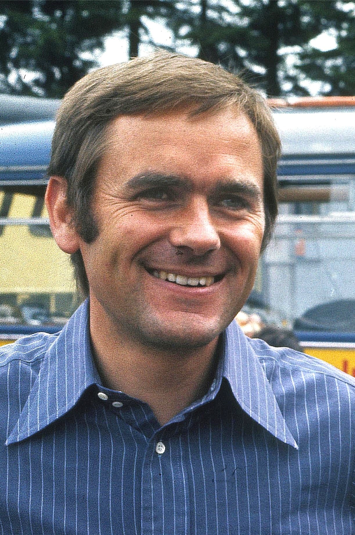 Head and shoulders of Jochen Neerpasch smiling, in a blue pinstriped shirt, at a race track.
