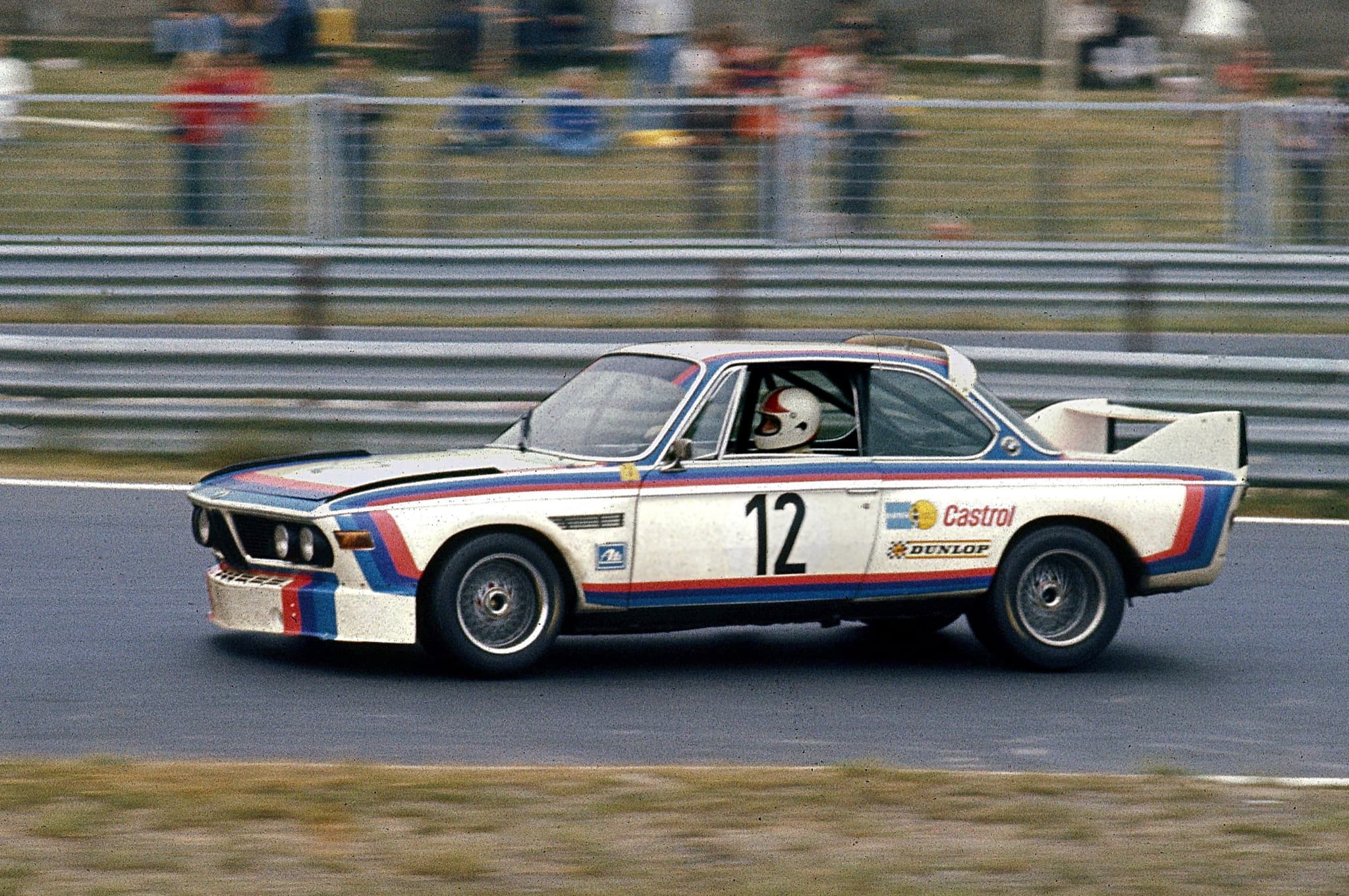 A white BMW 3.0 CSL racing car with BMW Motorsport stripes, number 12, panned on the Nürburgring straight.