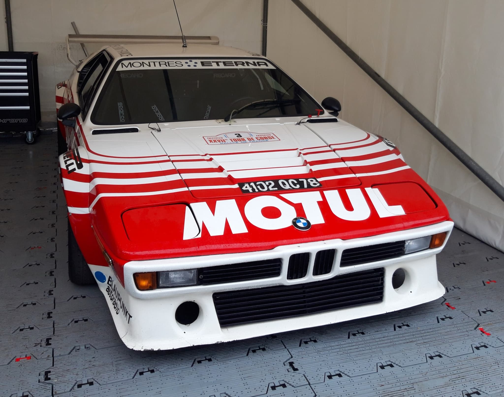 The red and white Motul rally BMW M1 nearly head-on in a tent, with a Tour de Corse rally plate on the bonnet. Opens a larger view