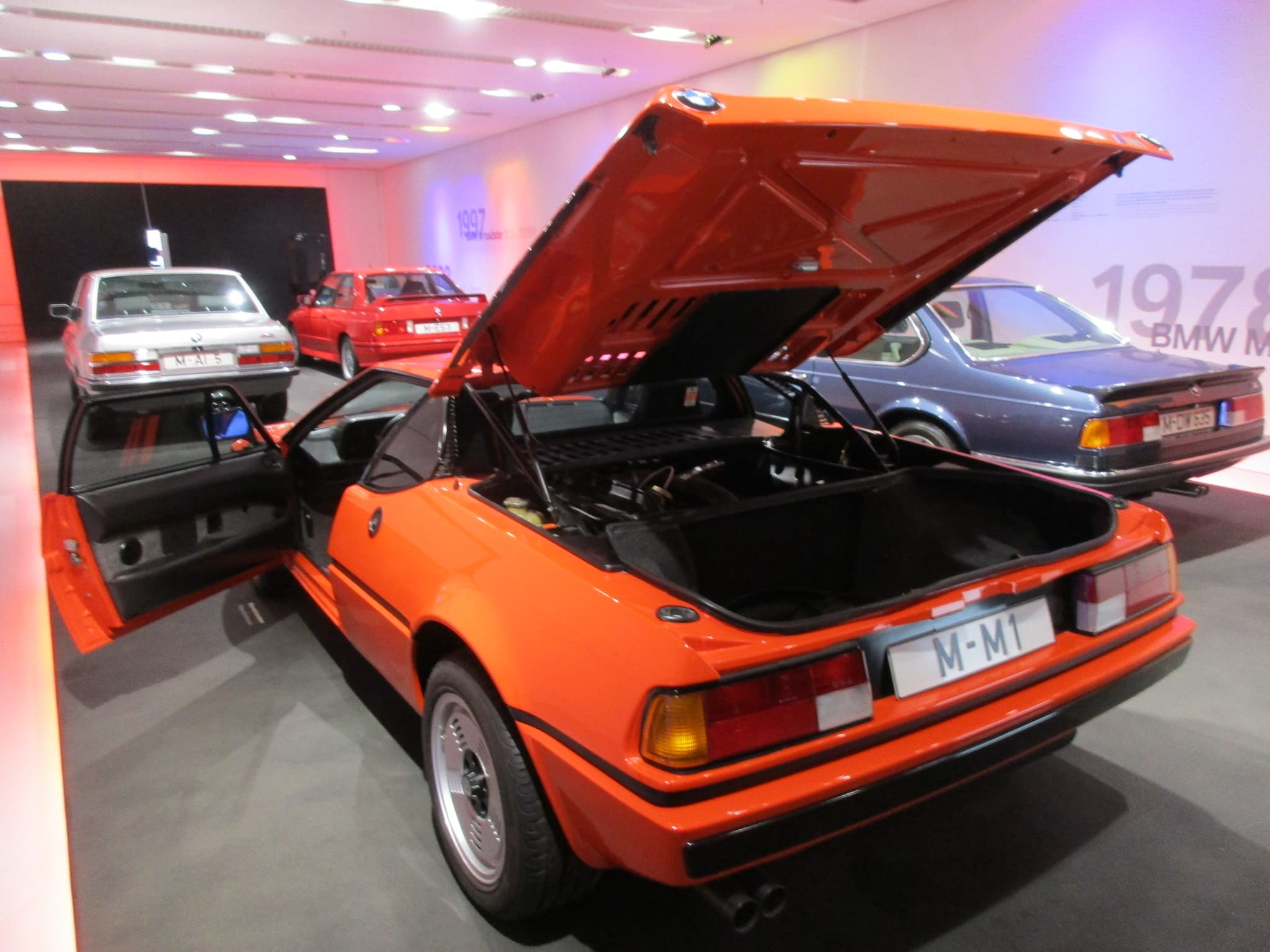 An orange BMW M1 with its whole rear deck raised and the driver's door open, and the carpeted luggage box behind the engine. Opens a larger view