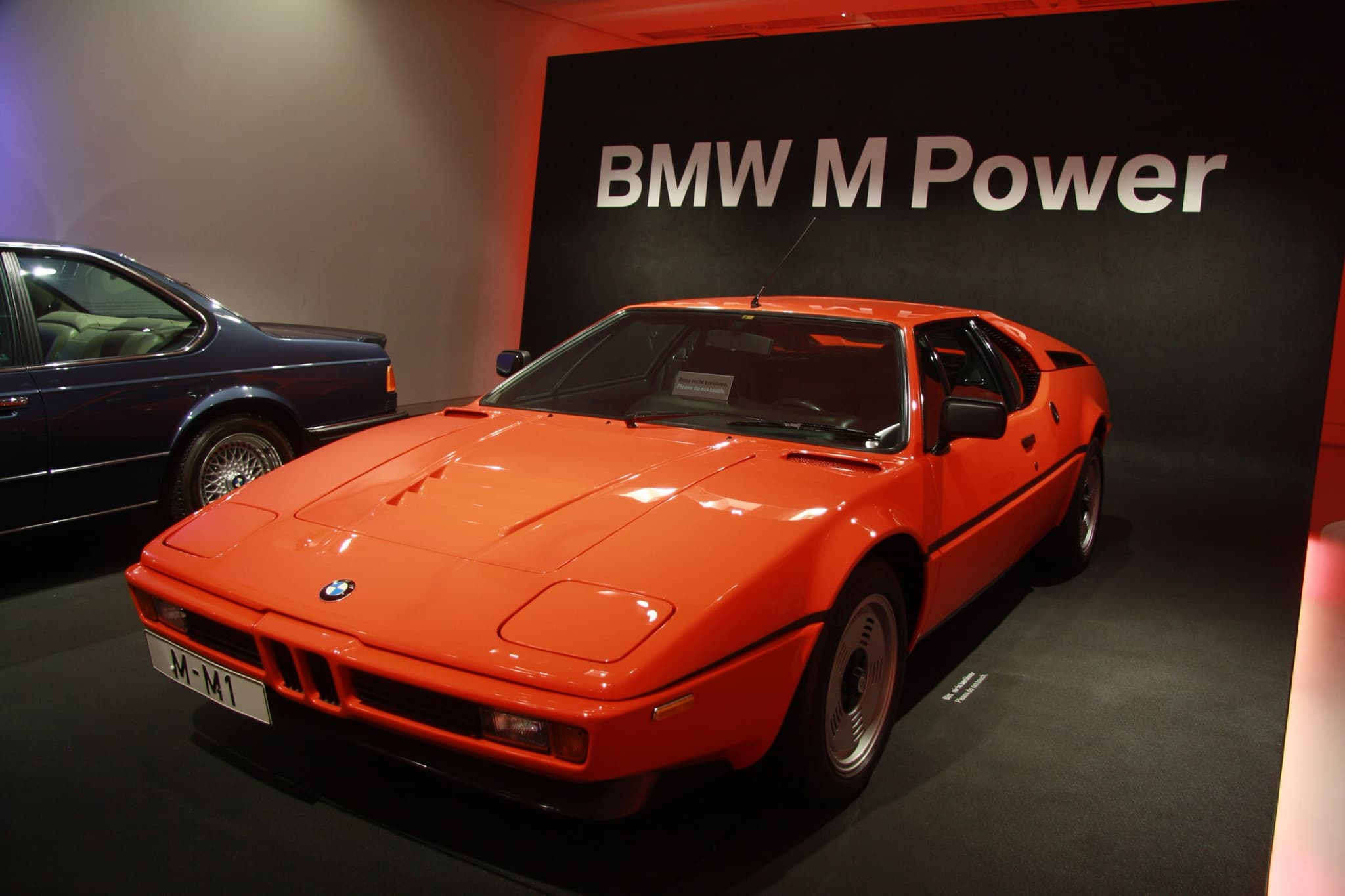 An orange BMW M1 in front of a wall lettered BMW M Power, front three-quarter, with another BMW coupé beside it.