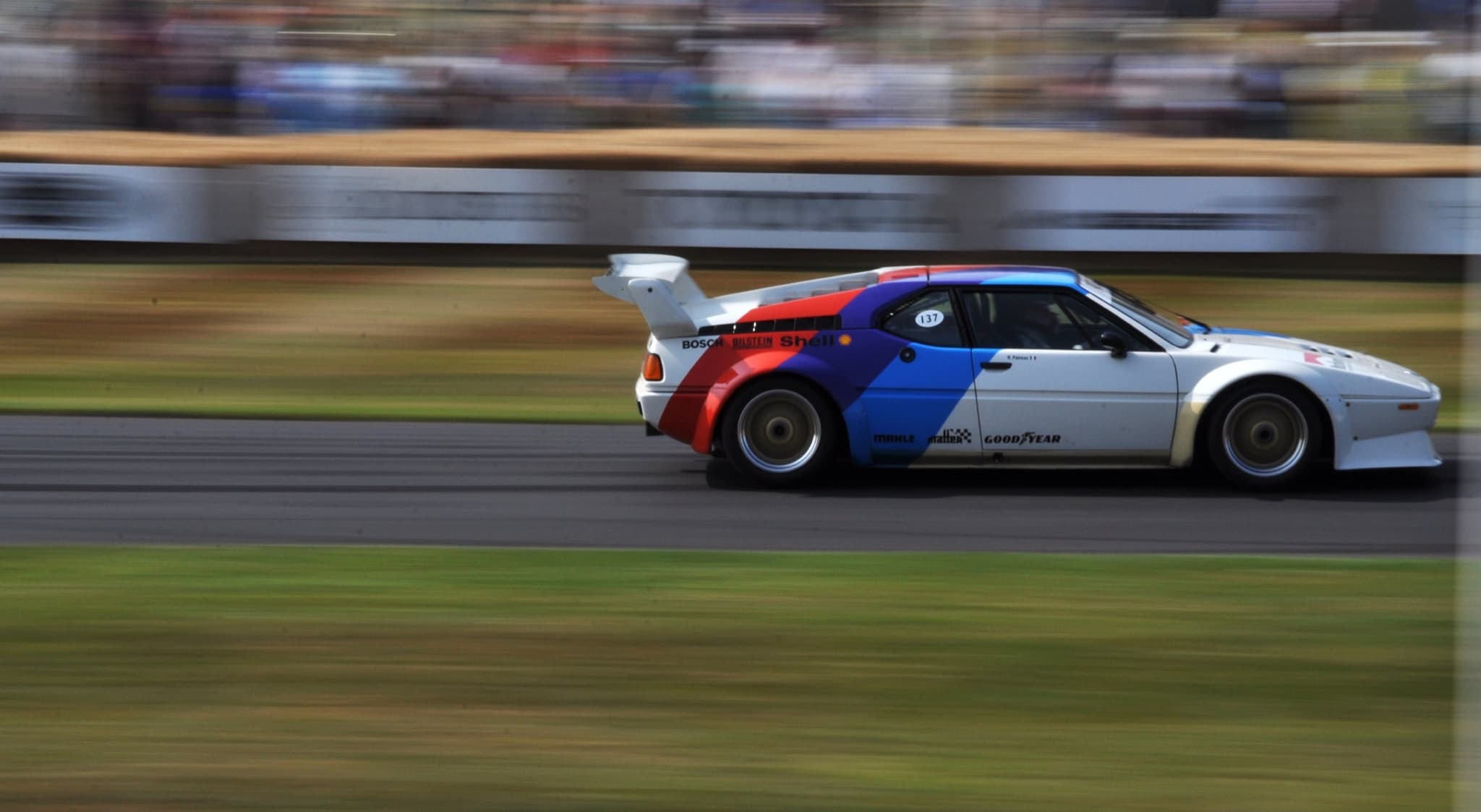 A white BMW M1 racing car with blue, violet and red stripes, flared wheel arches, a deep front spoiler and a tall rear wing, panned at speed past blurred straw bales and hoardings.