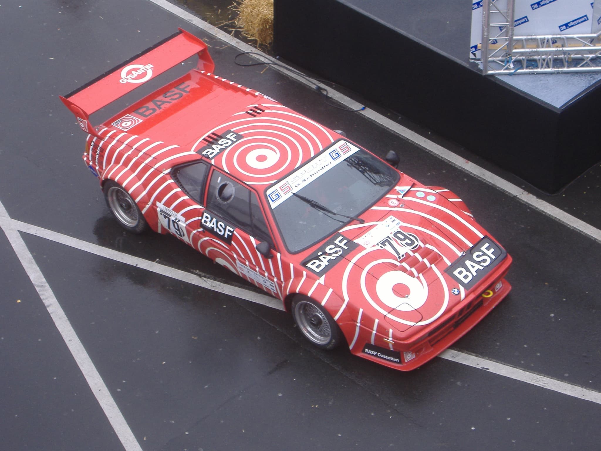 A racing BMW M1 in a red and white target scheme with BASF lettering and the number 79, seen from above on a wet paddock road.