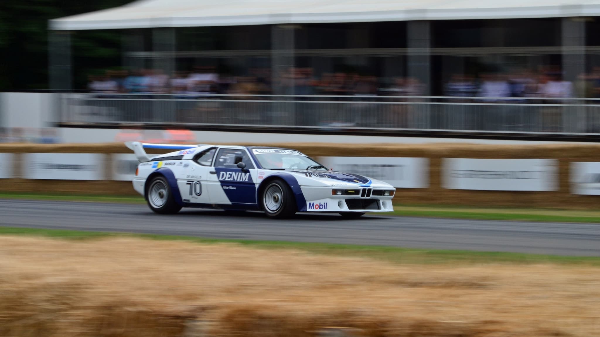 A white and dark blue BMW M1 Procar lettered DENIM and BMW ITALIA, number 70, panned past a grandstand.