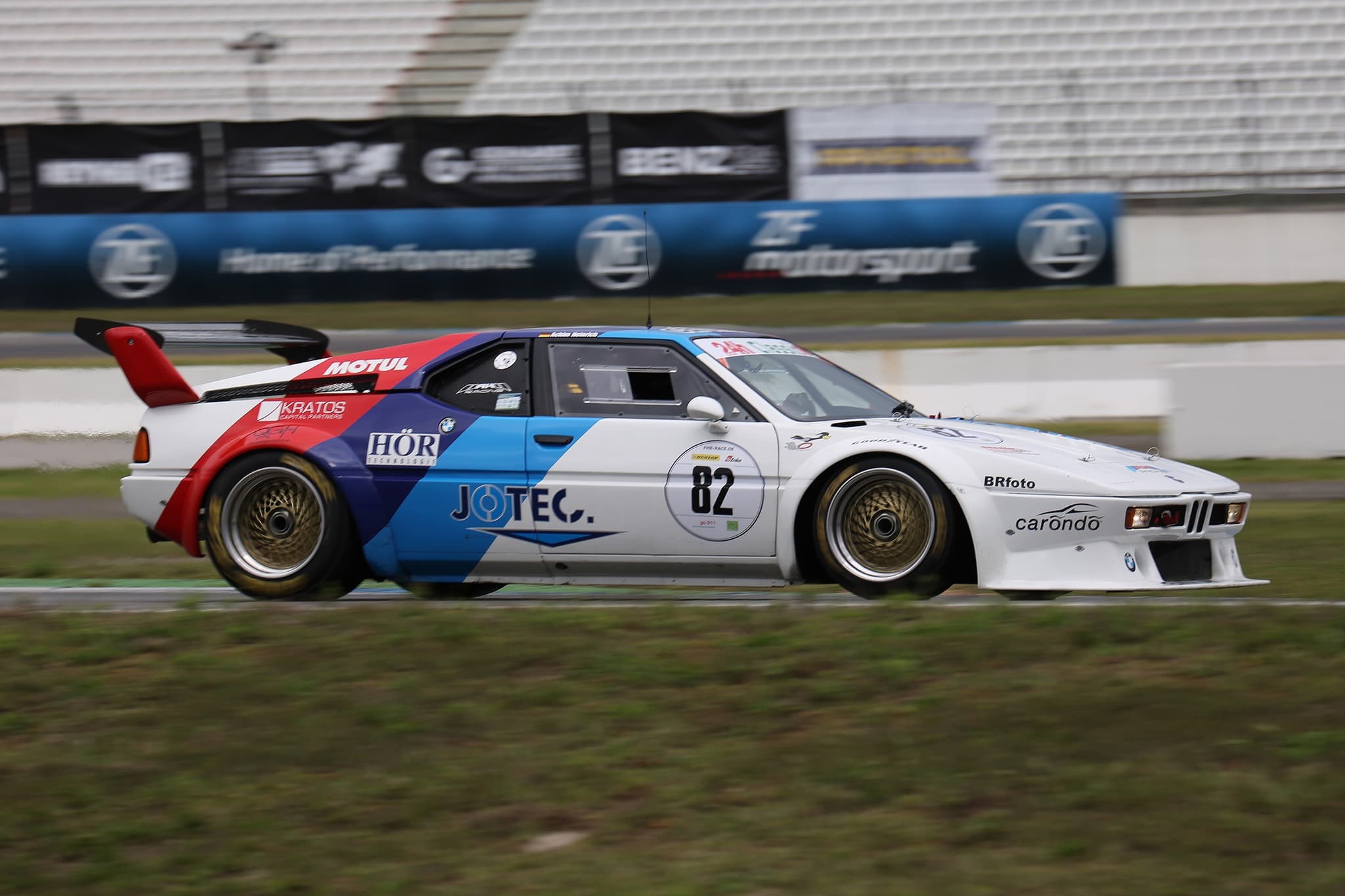 A white BMW M1 racing car with blue, violet and red stripes, sponsors' decals and gold wheels, number 82 on the door, panned at speed past circuit hoardings and an empty grandstand.